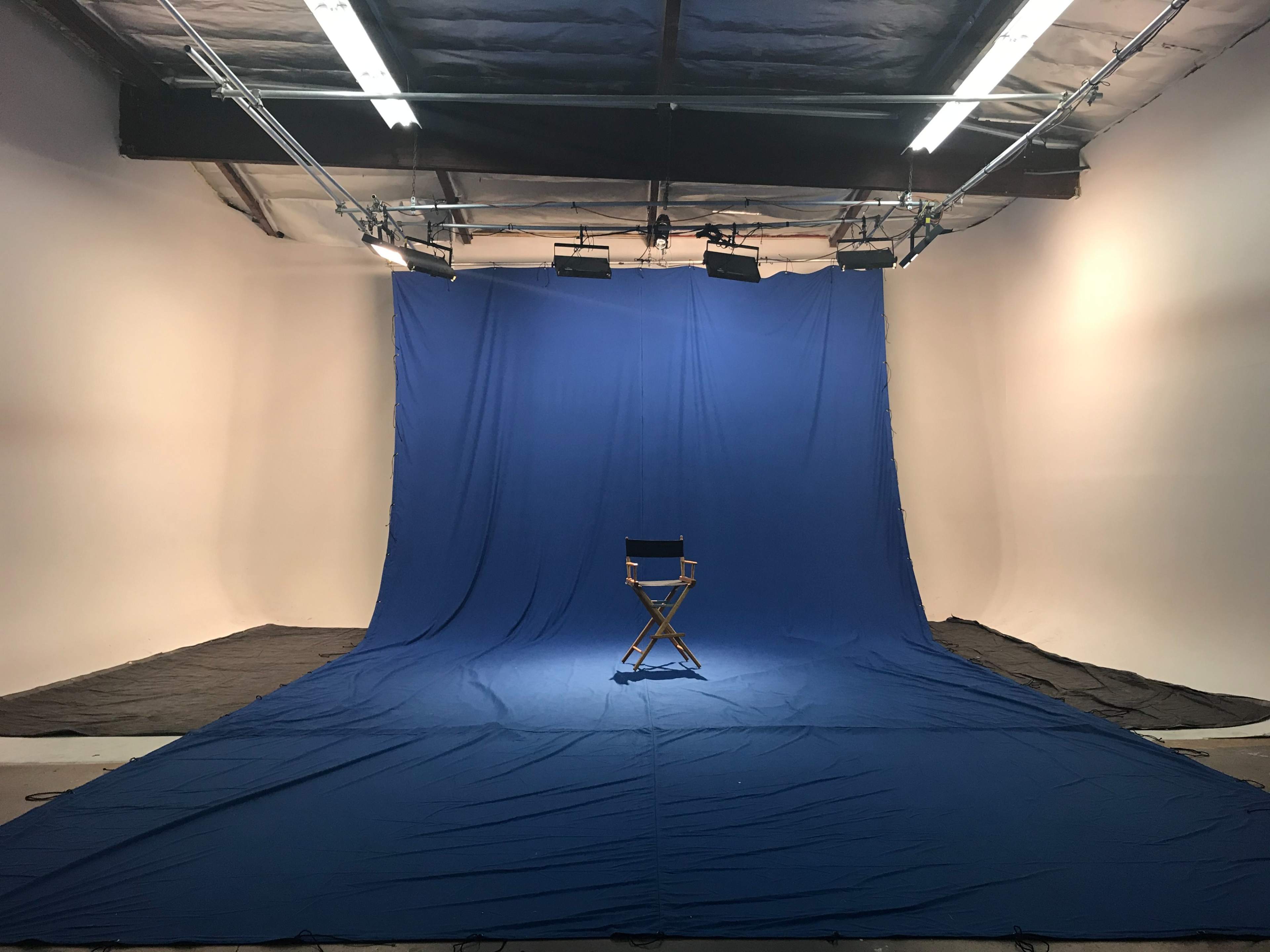 A blue backdrop covers the floor and walls of a studio, with a single director's chair positioned in the center under overhead lights.