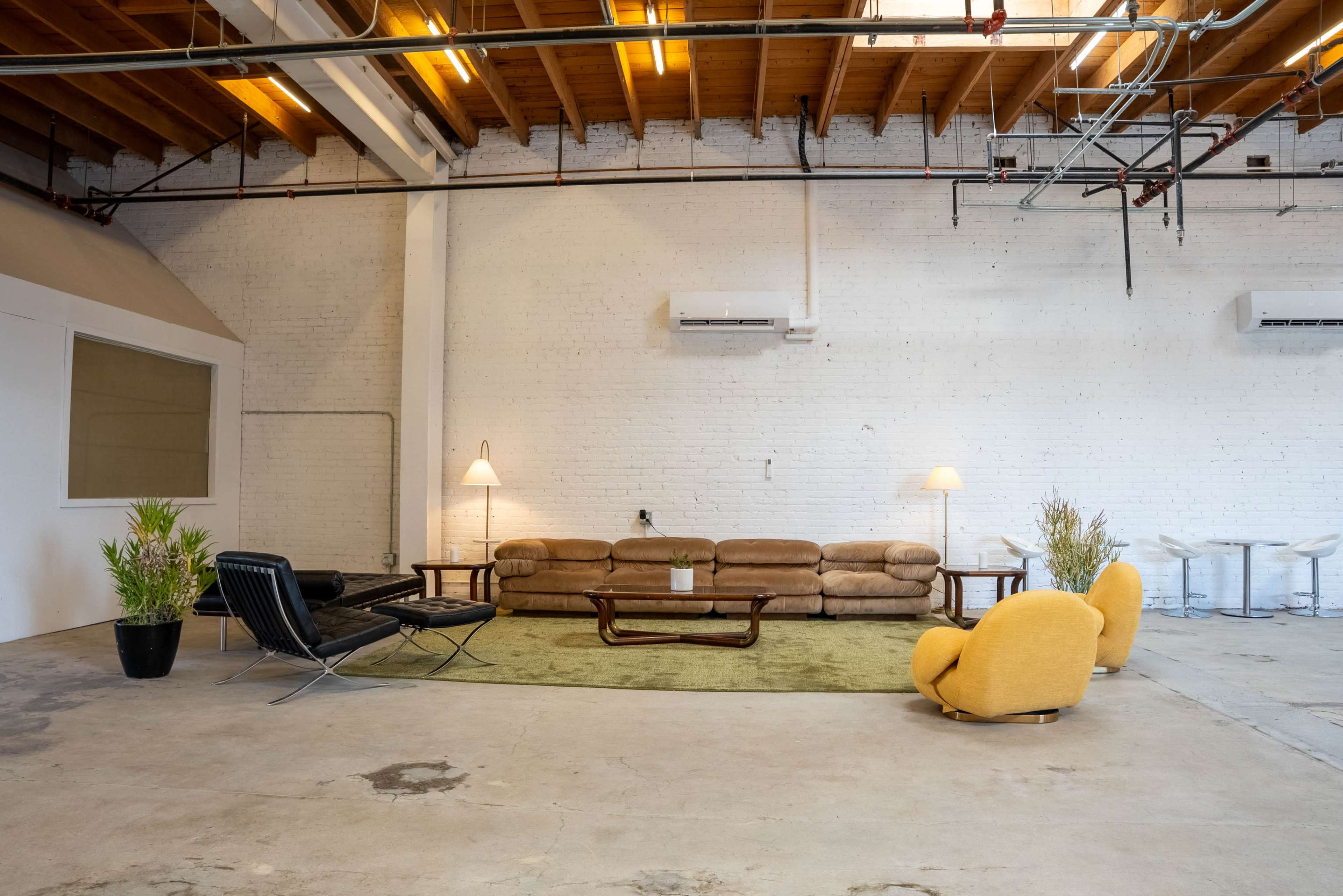 The image shows a spacious industrial-style room featuring a brown couch, a black lounge chair, yellow armchairs, and a green area rug, with exposed wooden beams and air conditioning units on the brick walls.