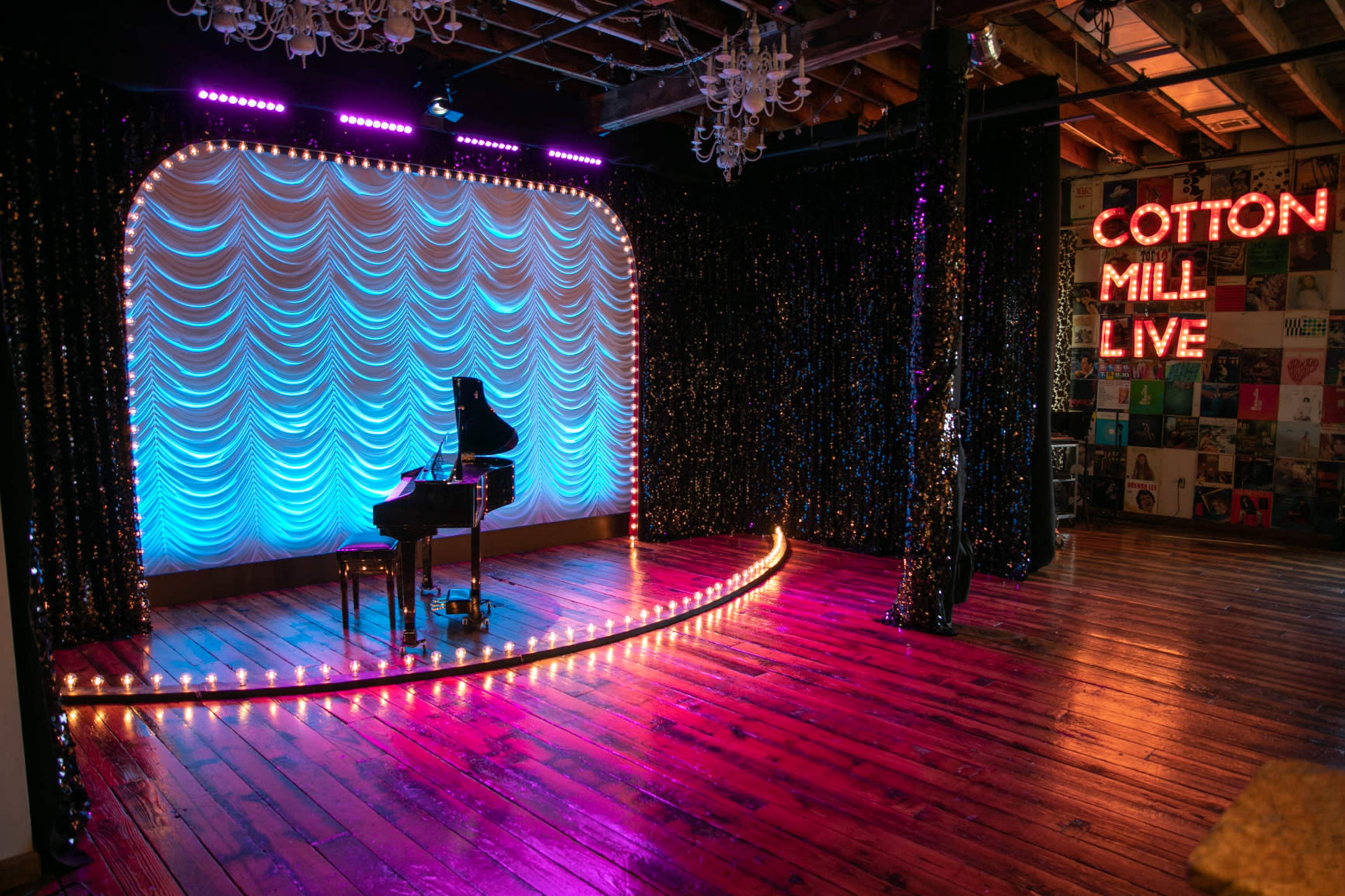 A performance stage with a grand piano, draped in soft blue fabric and surrounded by lights, located in a venue featuring a sign that reads "COTTON MILL LIVE."