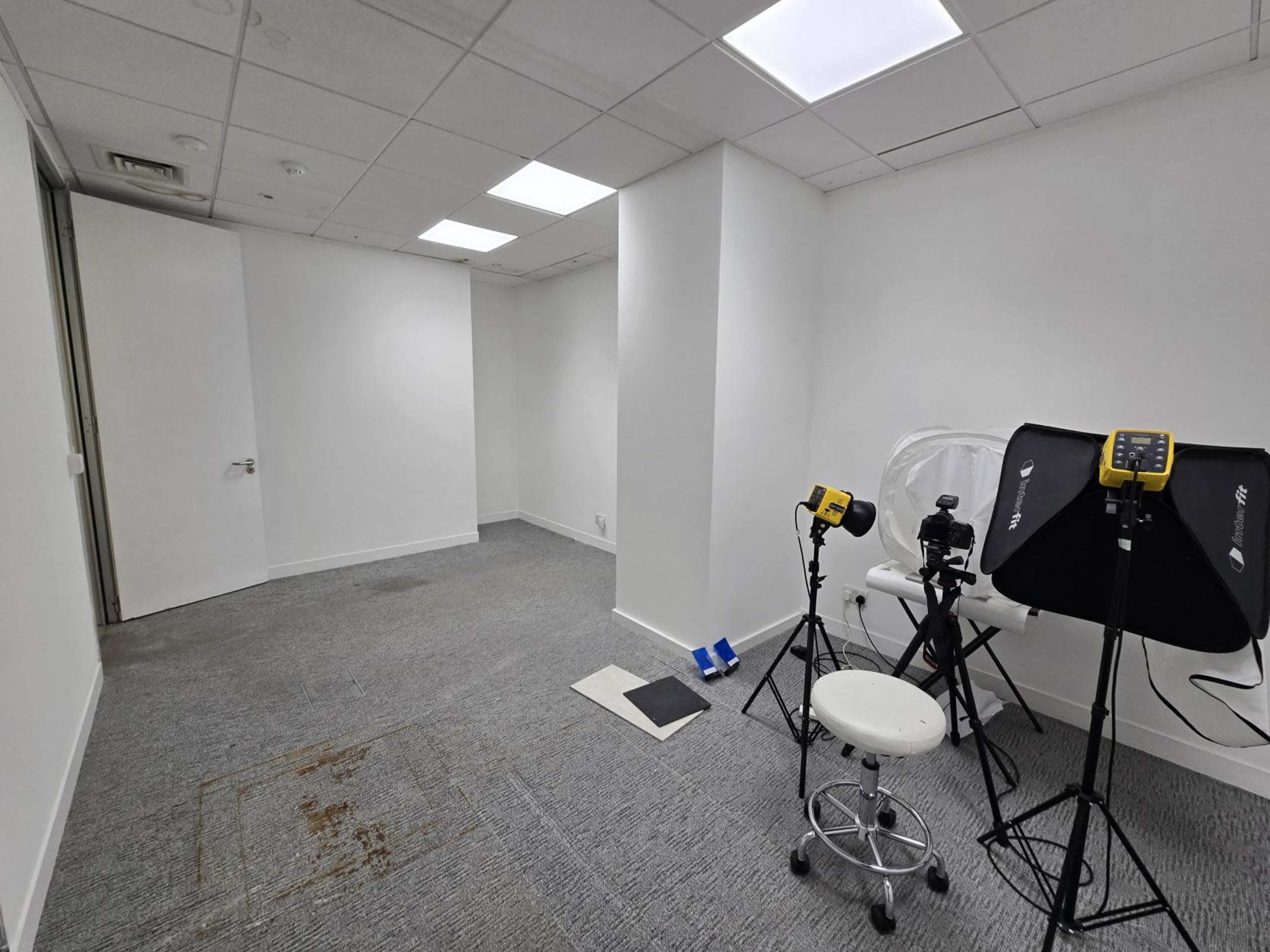 The image shows an empty room with gray carpet, white walls, and photography equipment including lights and a stool.