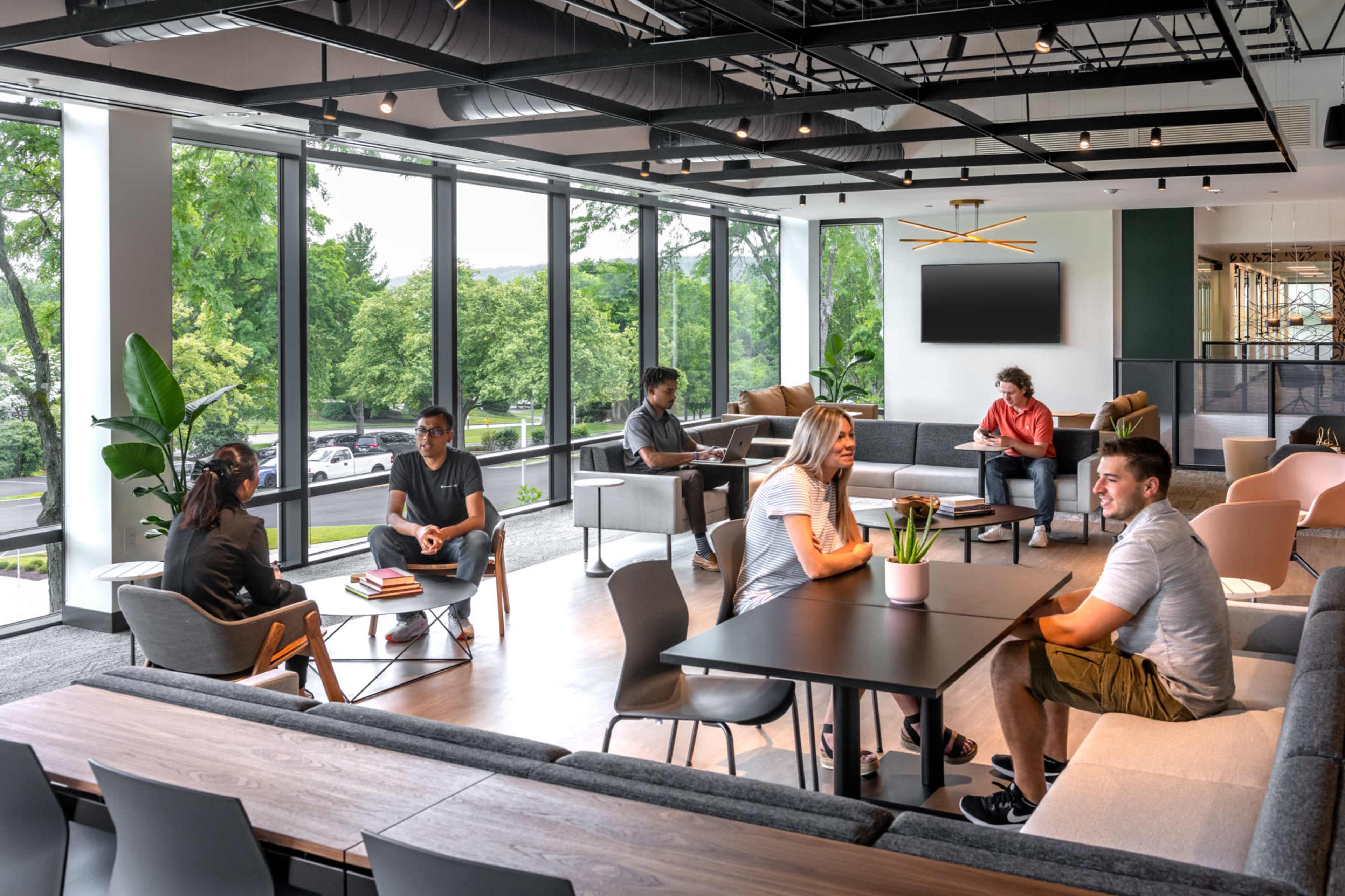 A modern workspace features groups of people engaged in conversations and activities around various seating arrangements and tables near large windows.
