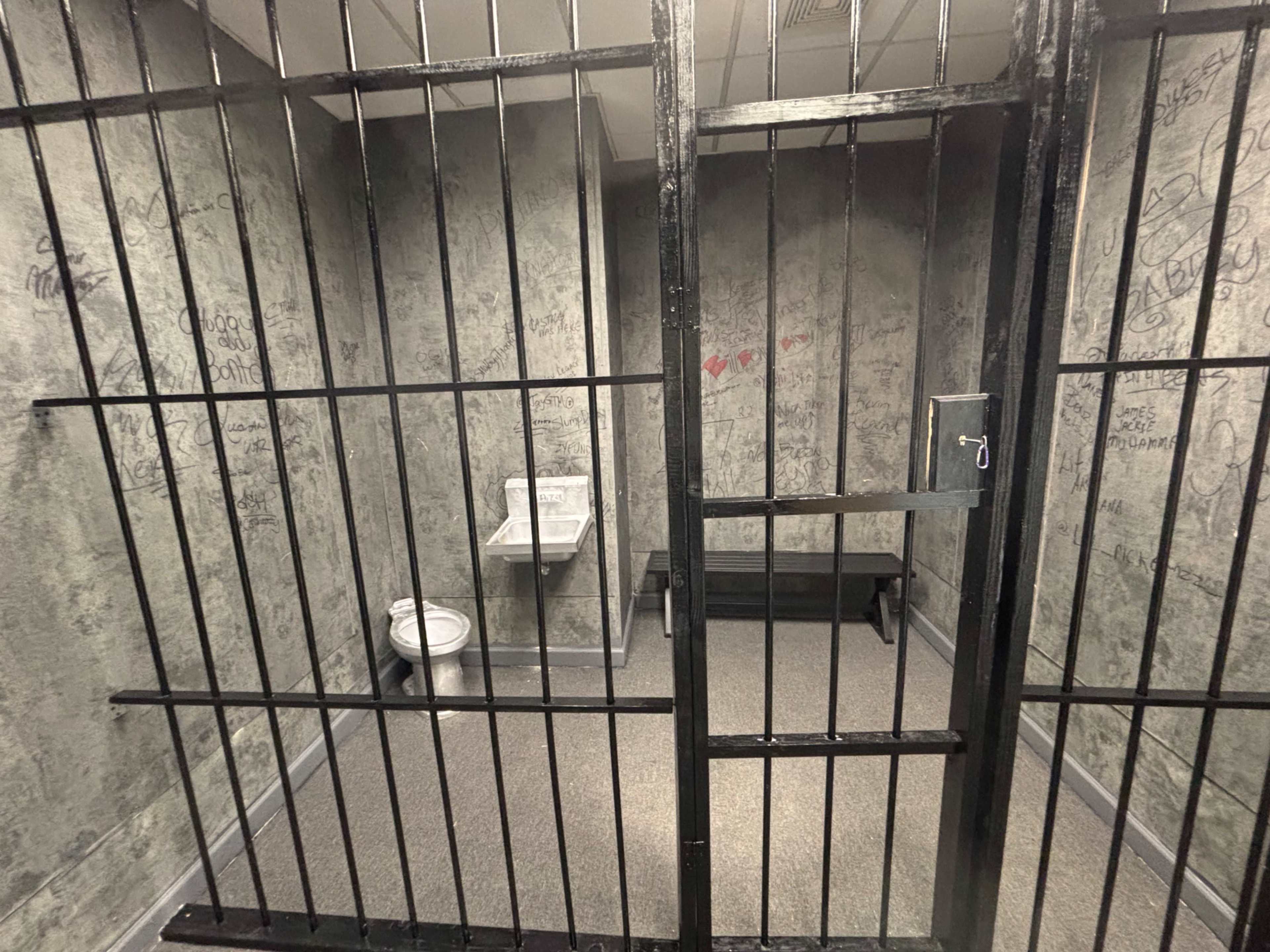 The image shows an empty jail cell with metal bars, a toilet, and a bench, featuring graffitied walls.