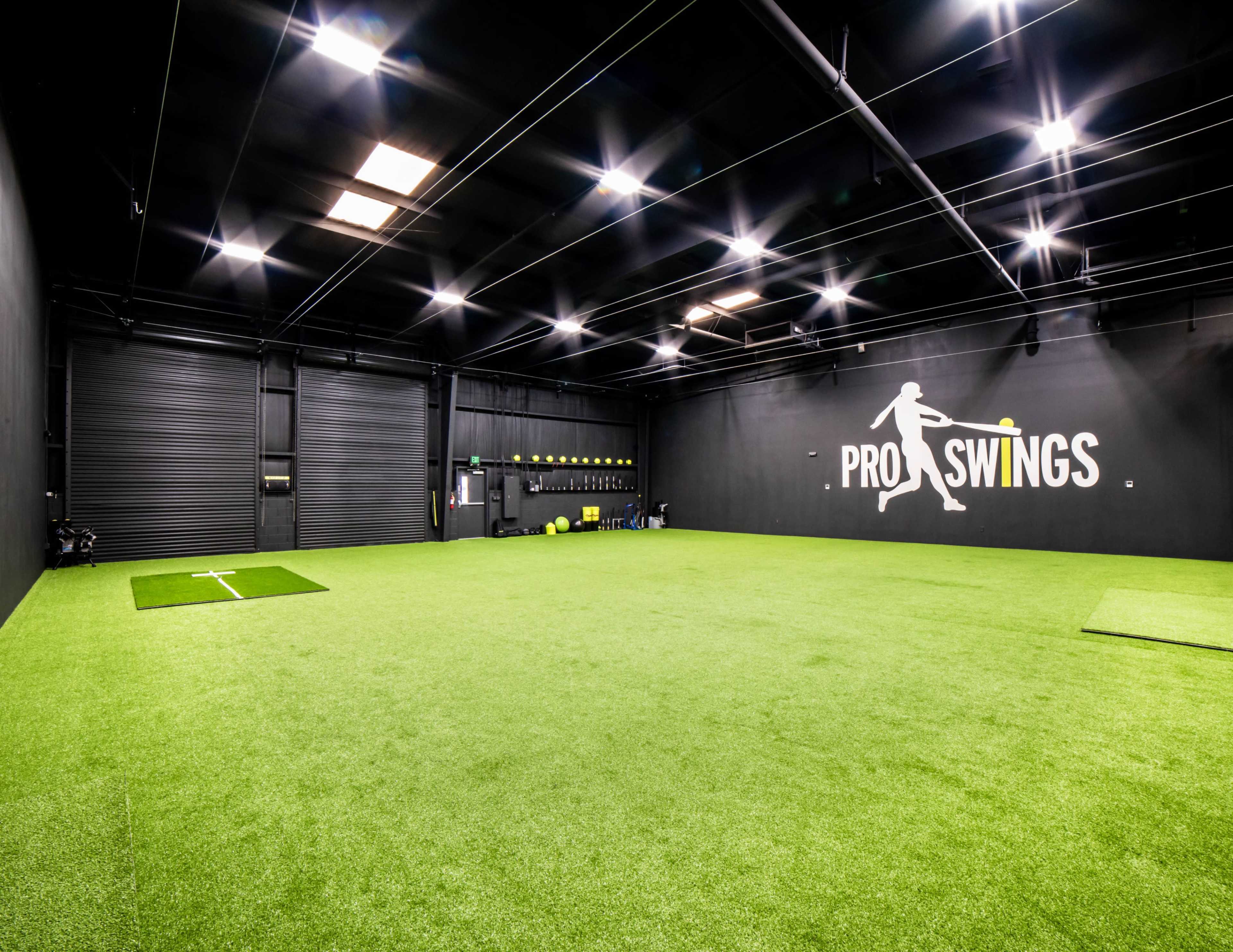 The image shows a large indoor space with a synthetic green turf floor, equipped for sports practice, featuring a black wall with the text "PRO SWINGS."