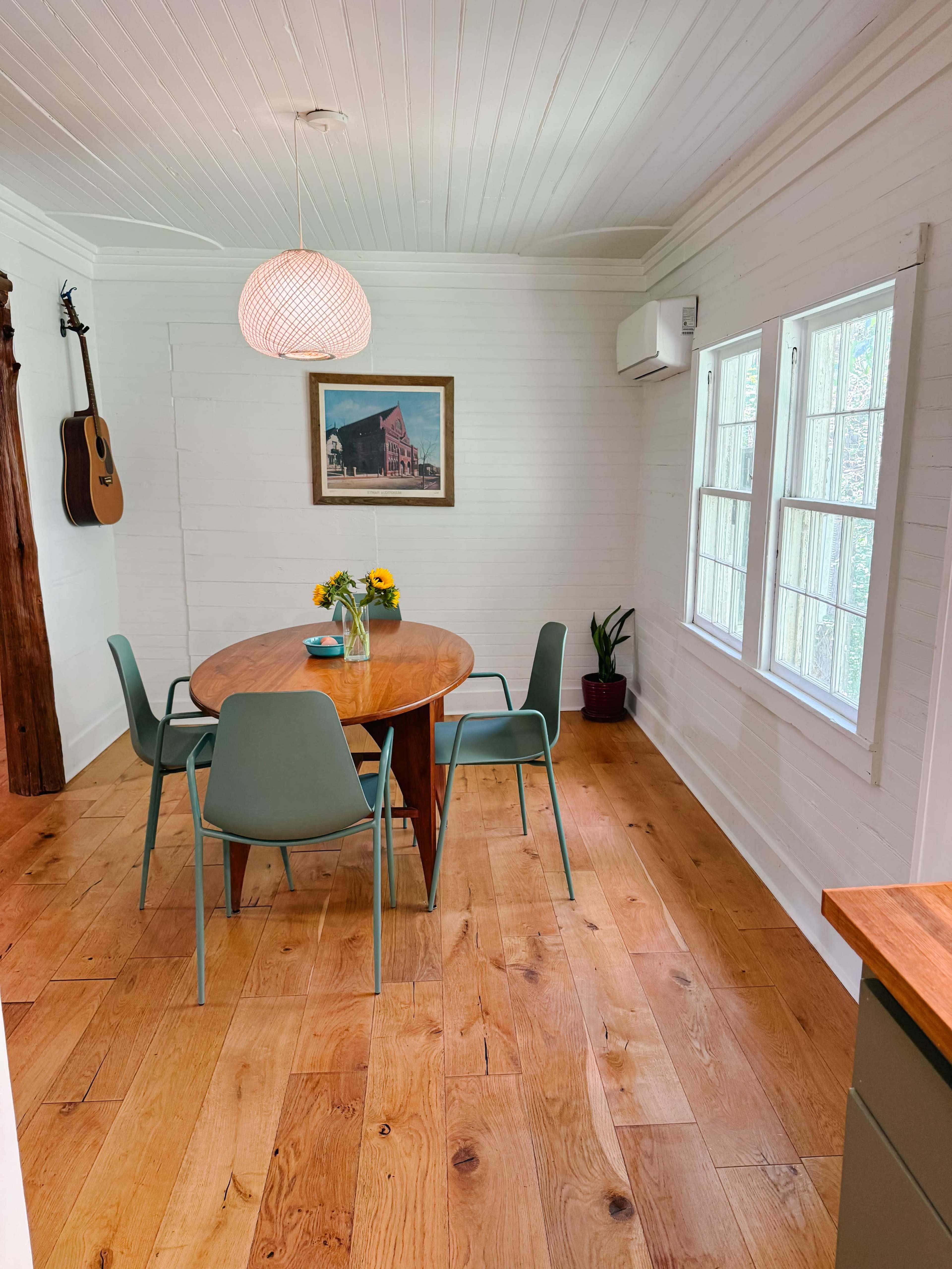 The image depicts a bright dining area featuring a round wooden table surrounded by four chairs, a vase of sunflowers, and a framed picture on the wall, with a guitar hanging nearby.