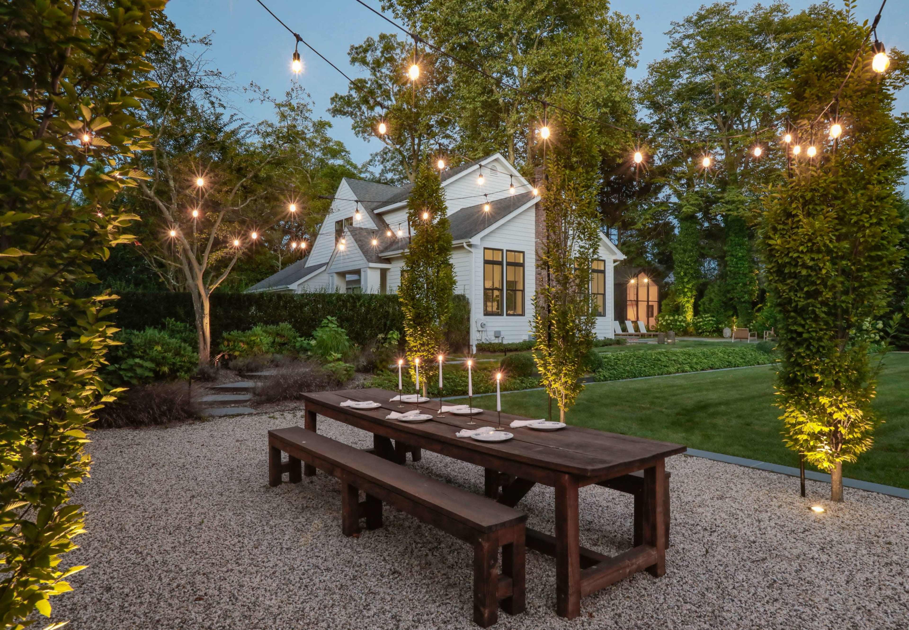 A long wooden dining table with candles is set in a gravel patio surrounded by trees and a well-kept lawn, illuminated by string lights overhead.