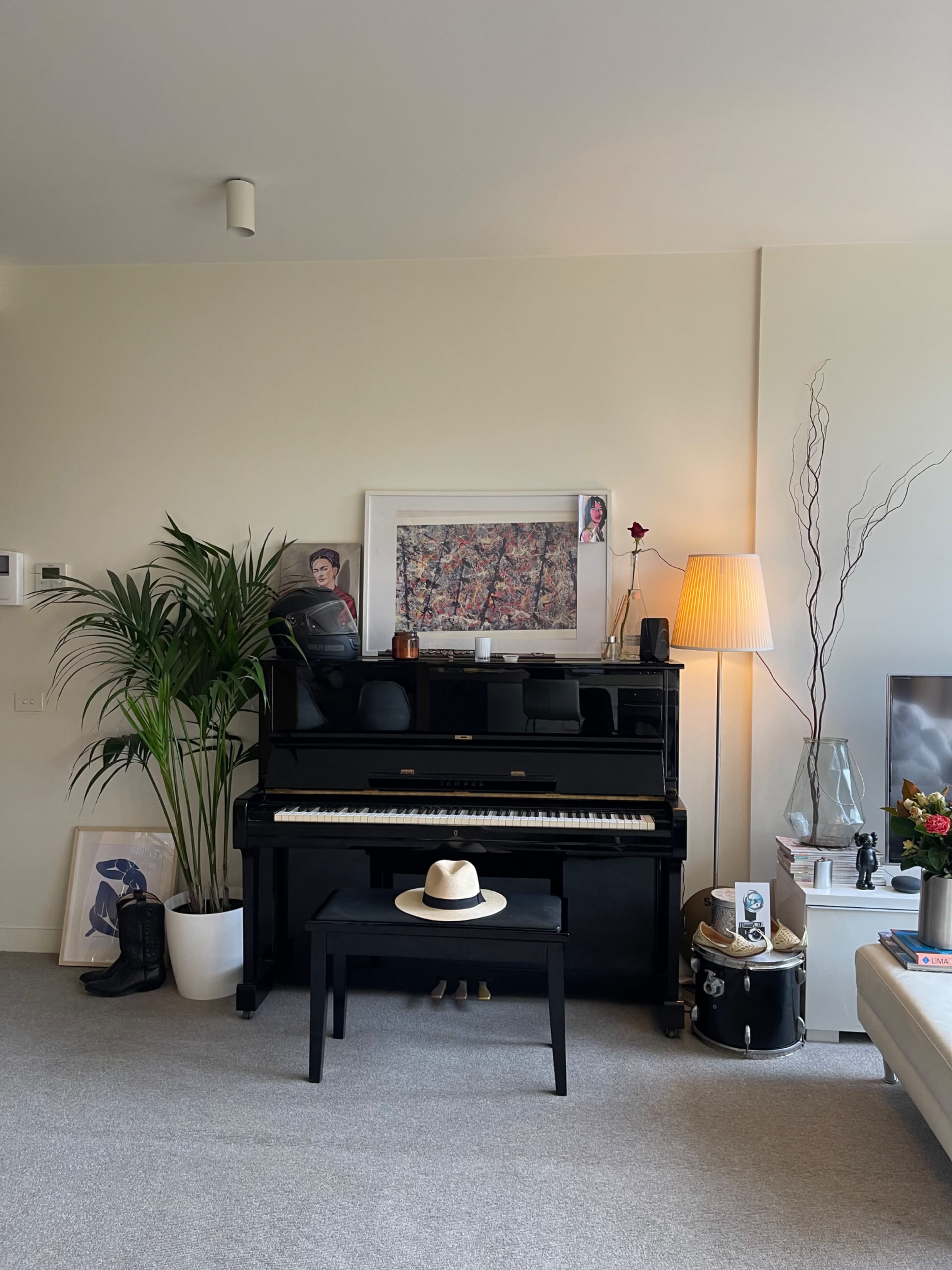 A living room with a black piano, a wooden bench, a hat on the bench, various plants, and artwork on the walls.