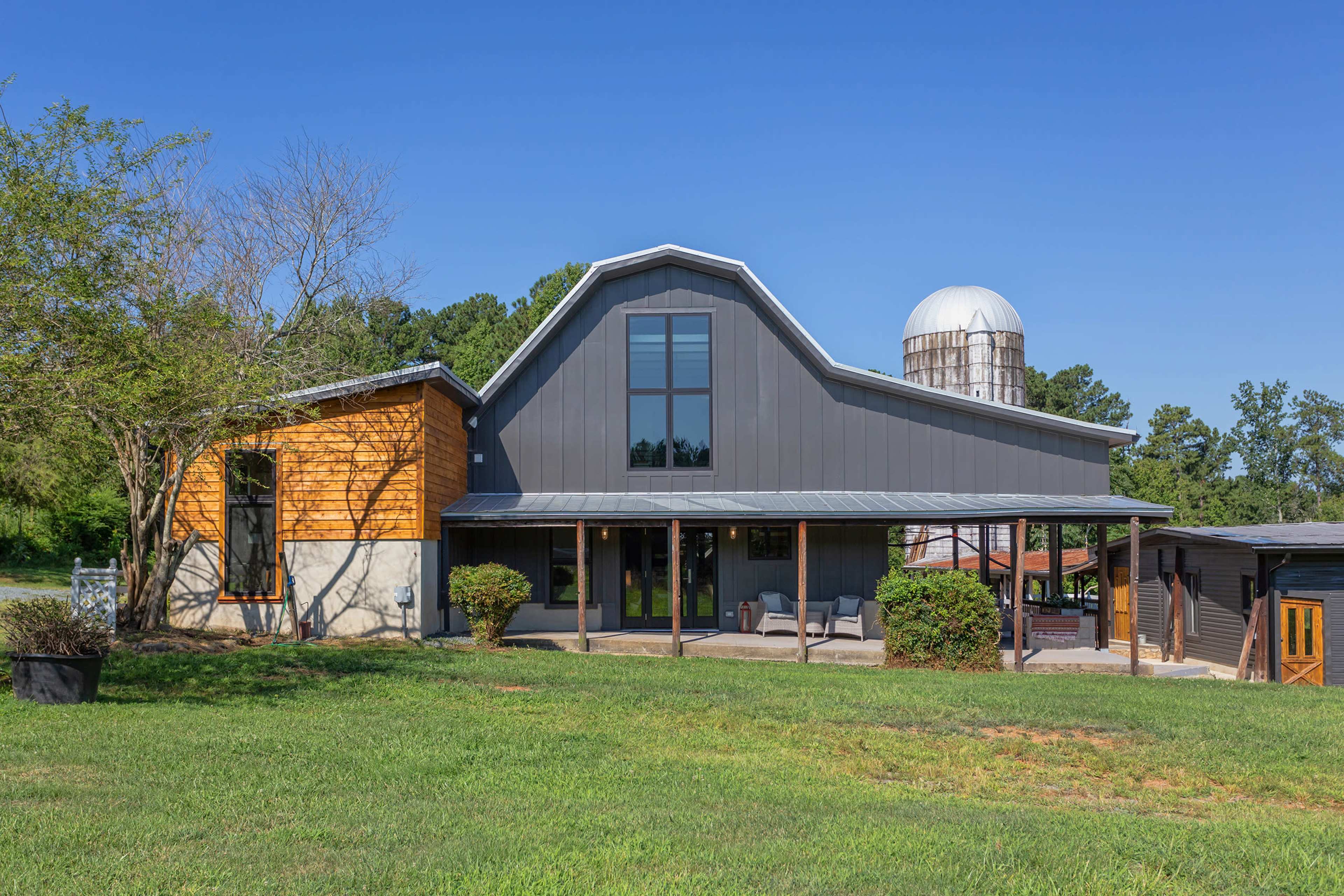 A modern farmhouse with a combination of wooden and metal siding, featuring a large window and an adjoining silo, sits in a grassy yard.