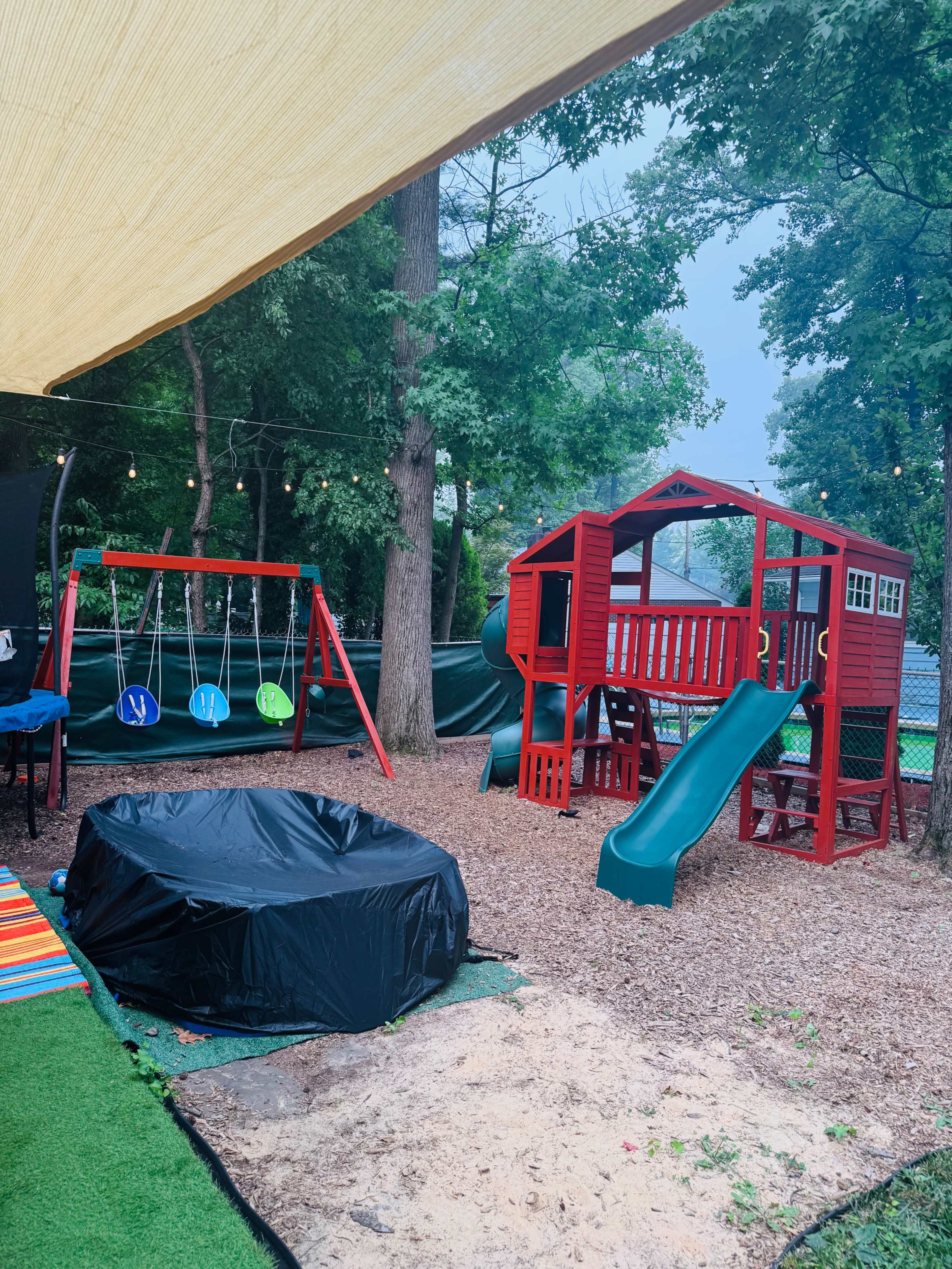 The scene features a backyard playground with a red playset that includes a slide and a wooden fort, alongside a swing set with green swings and a black covered fire pit area.