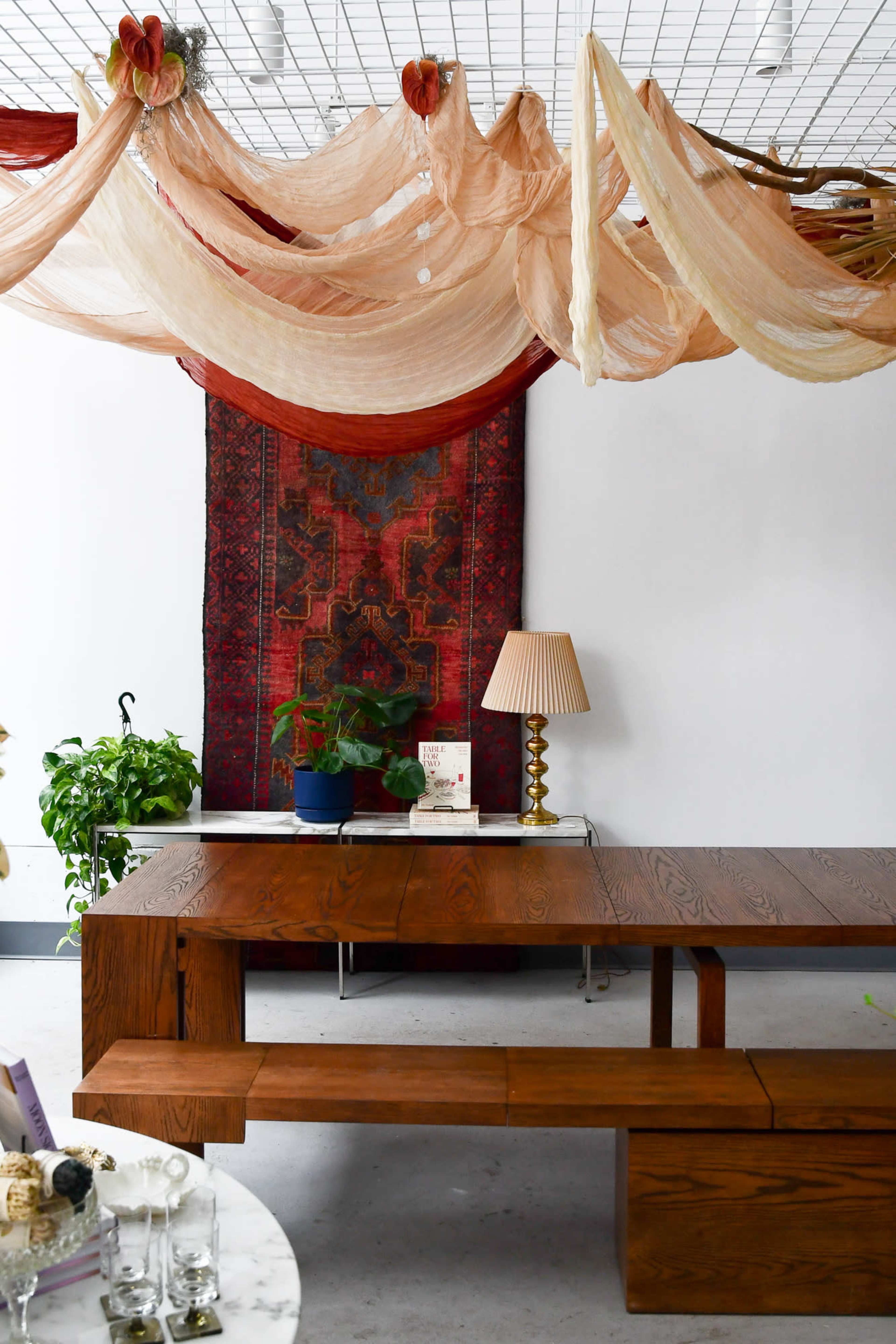 A wooden table with a long bench is set underneath draped fabric and a decorative rug hanging on the wall, alongside a lamp and potted plant.