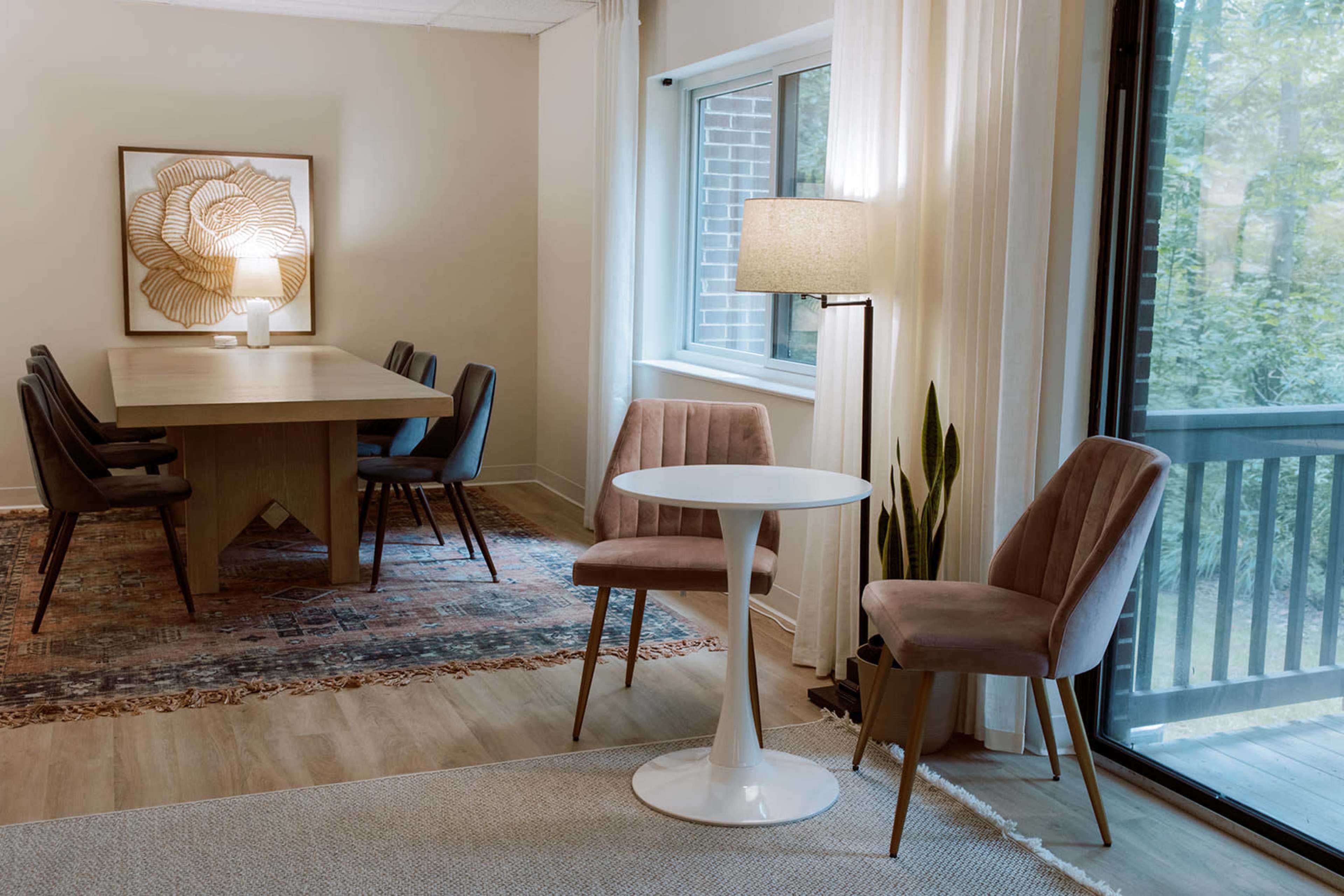 The image shows a modern dining area with a large wooden table, upholstered dining chairs, a small round table, and a floor lamp, all adjacent to a large window with a view of greenery.