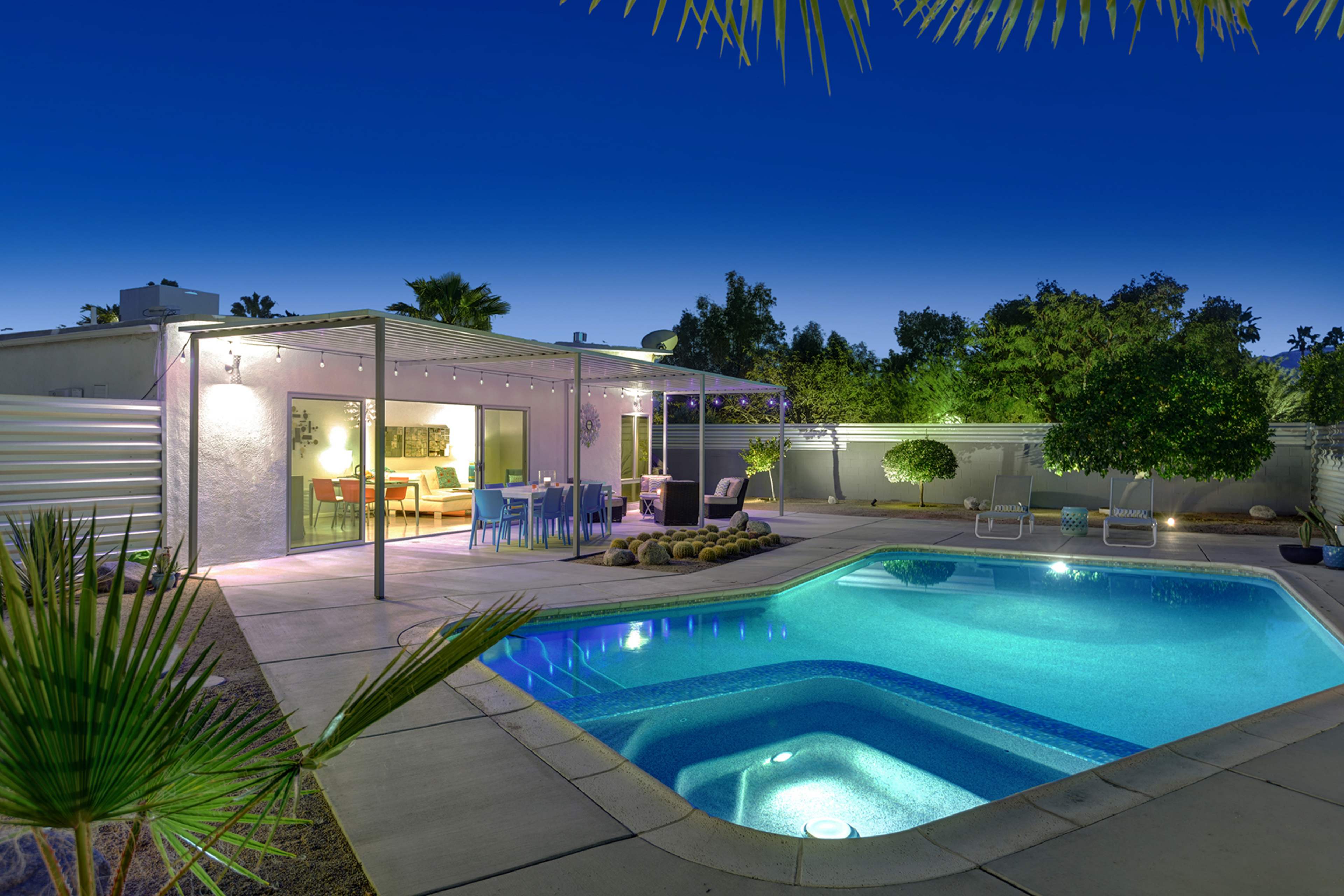 The scene features a modern backyard with a illuminated pool, a covered patio with dining furniture, and surrounding greenery under a clear evening sky.