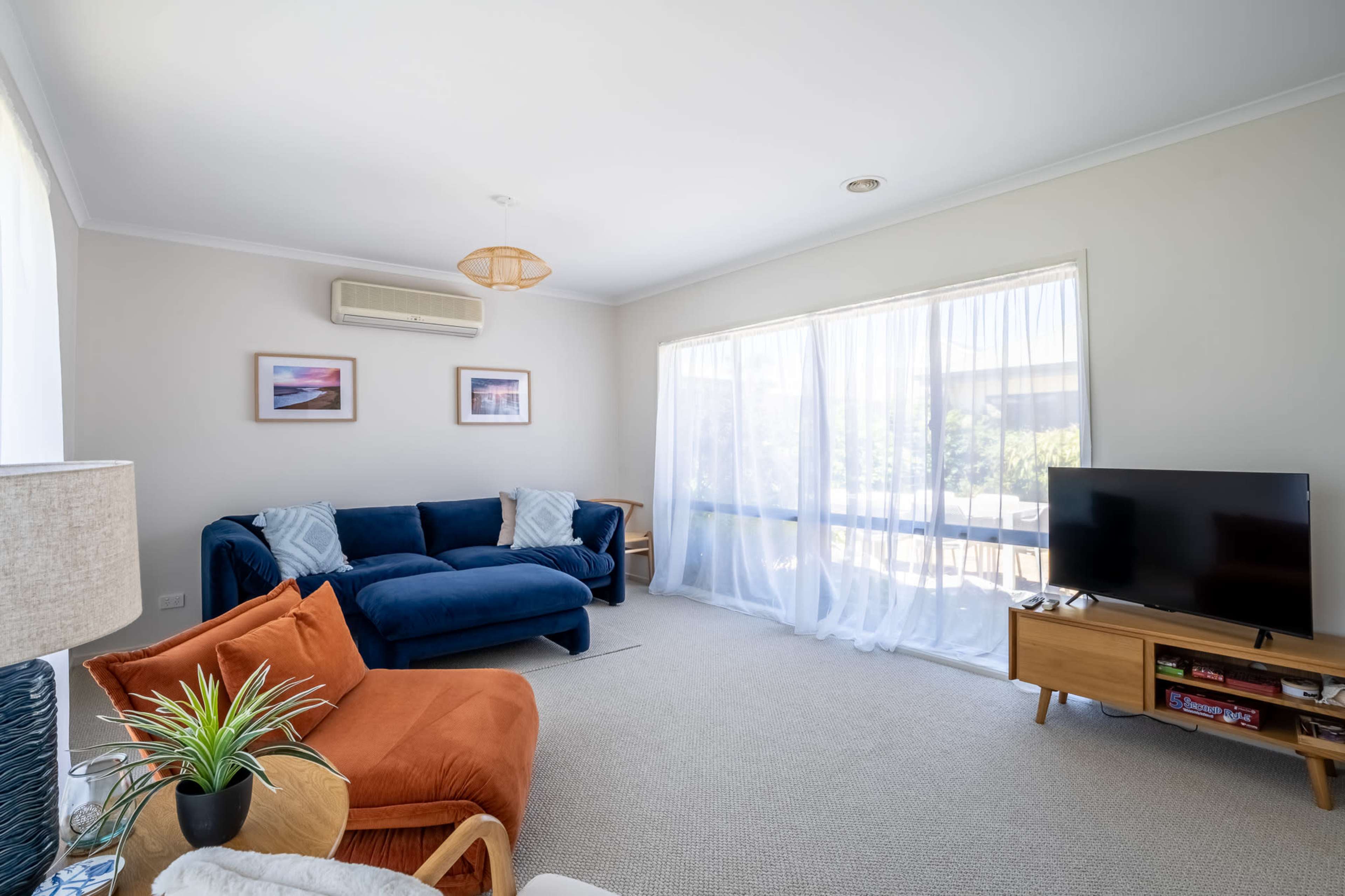 The living room features a blue sectional sofa, an orange accent chair, a television on a wooden stand, and large windows covered with light, sheer curtains.