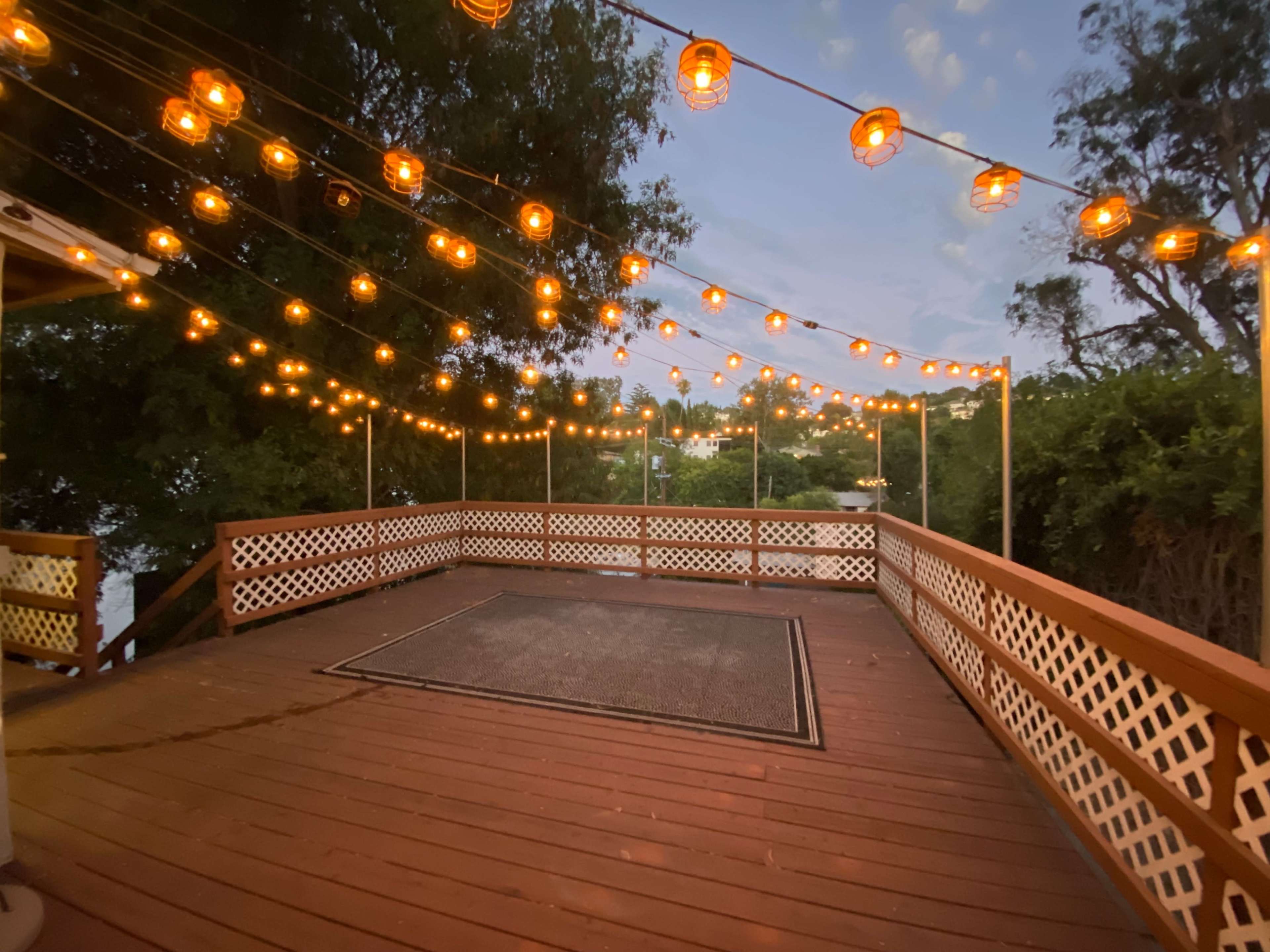 A wooden deck is illuminated by string lights, surrounded by greenery and overlooking a body of water at dusk.