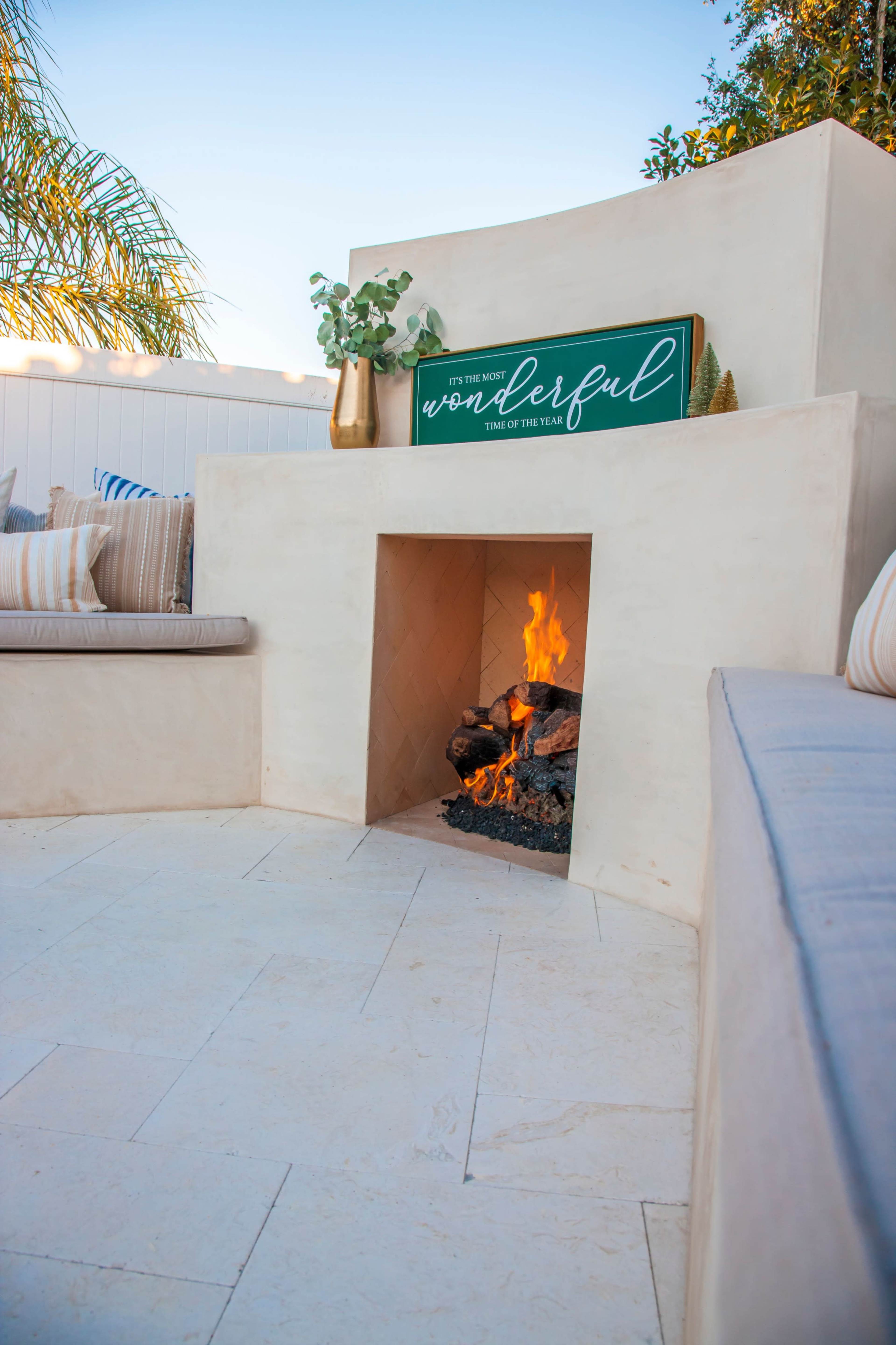 A modern outdoor fireplace is set into a smooth, light-colored structure, surrounded by cushions and a decorative sign that reads "It's the most wonderful time of the year," while a small fire crackles inside the hearth.
