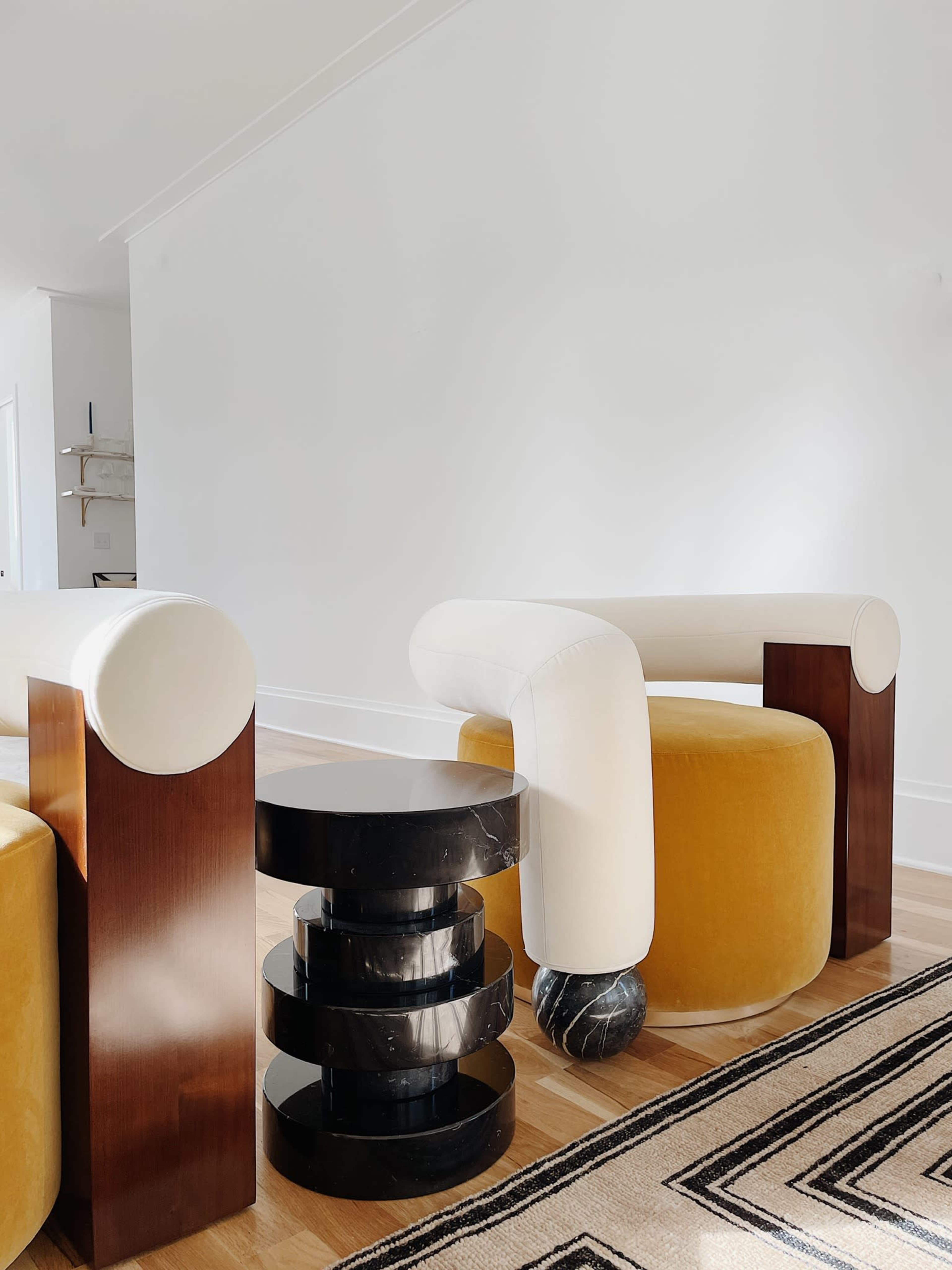 A modern living space featuring a white armchair with cylindrical wooden arms, a yellow pouf, and a black stacked side table against a white wall.