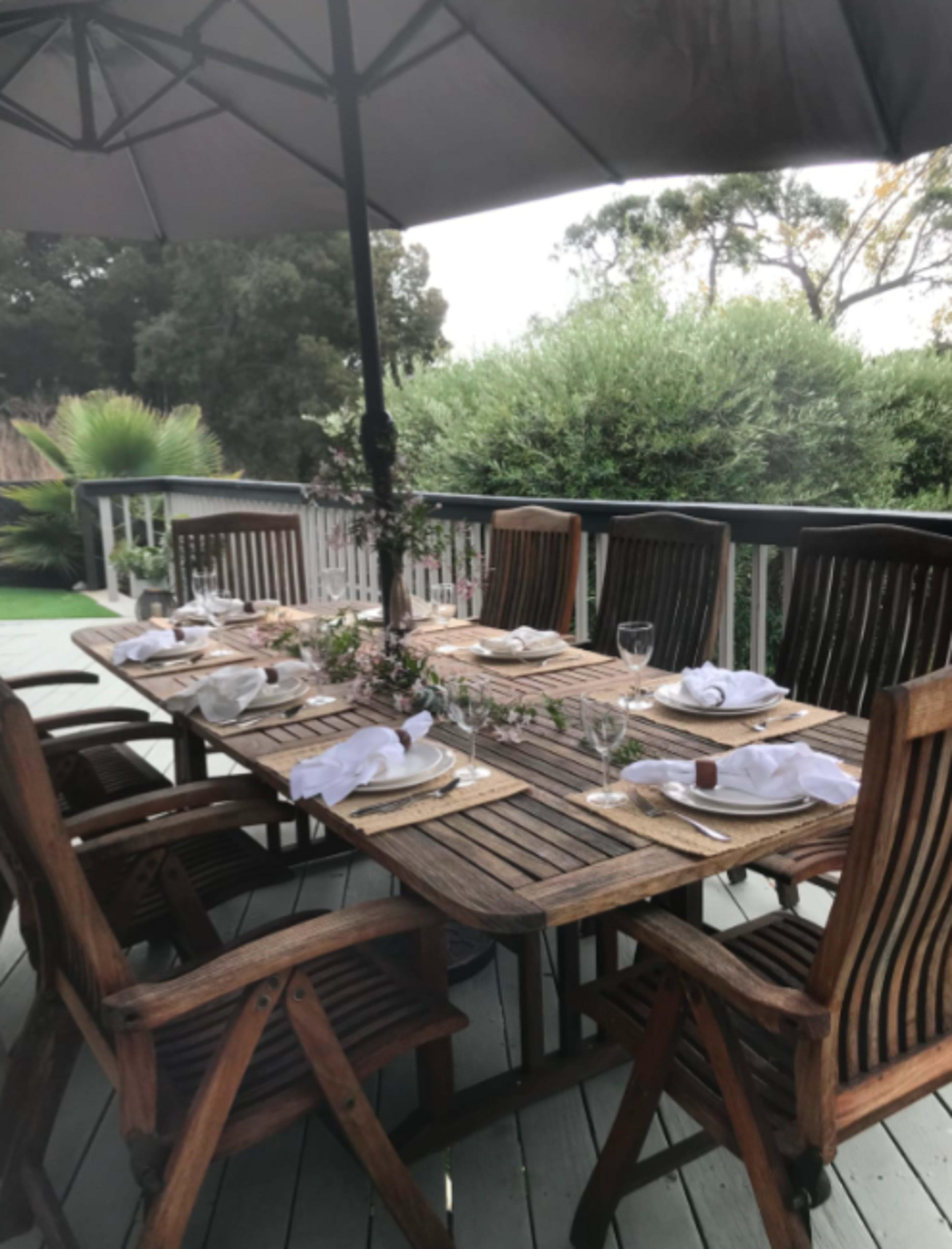 A wooden outdoor dining table is set with plates and glasses under a large umbrella on a deck surrounded by greenery.