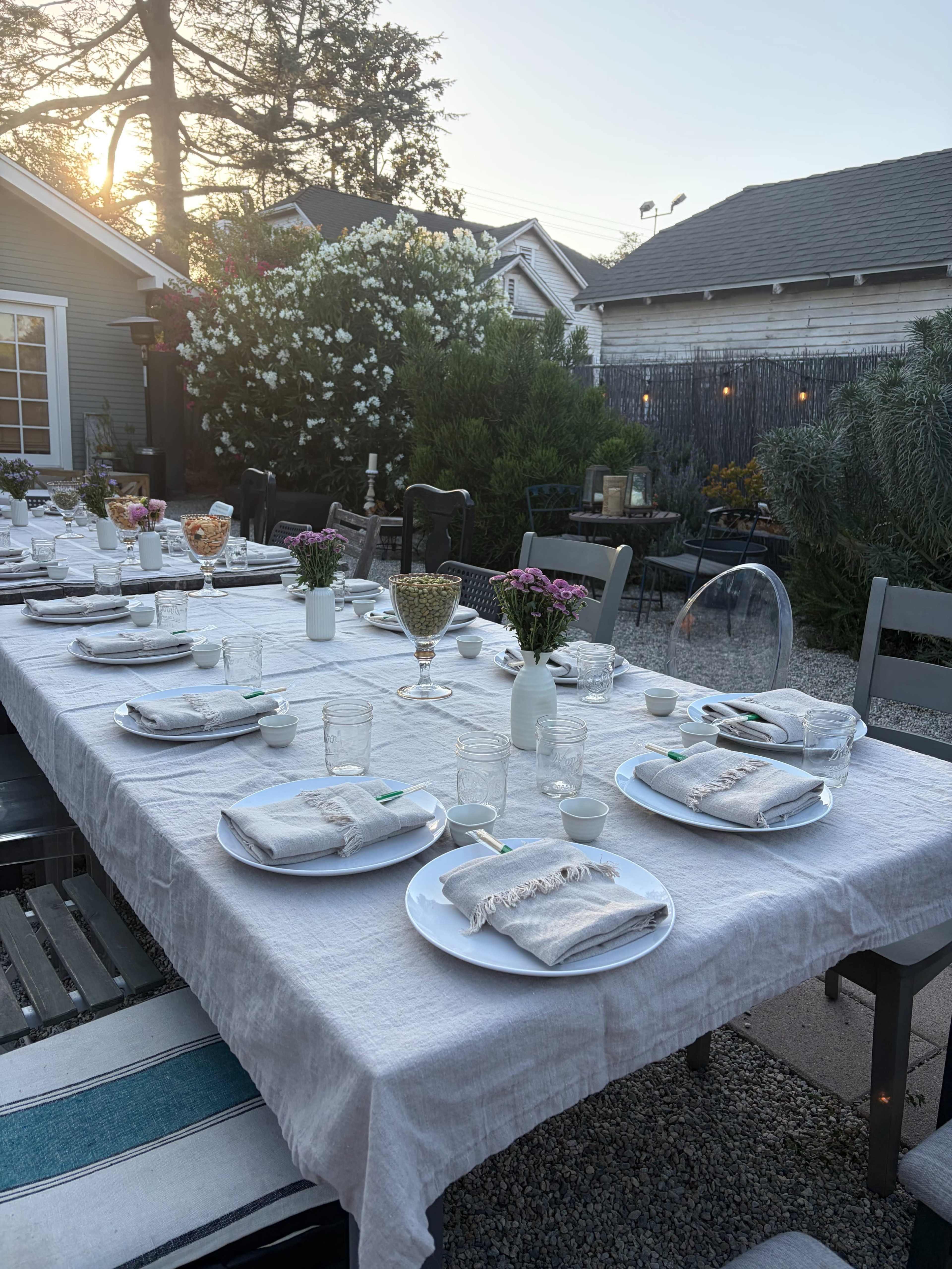 A long outdoor dining table is set with plates, glasses, and decorative flowers, surrounded by greenery and bathed in warm sunlight.