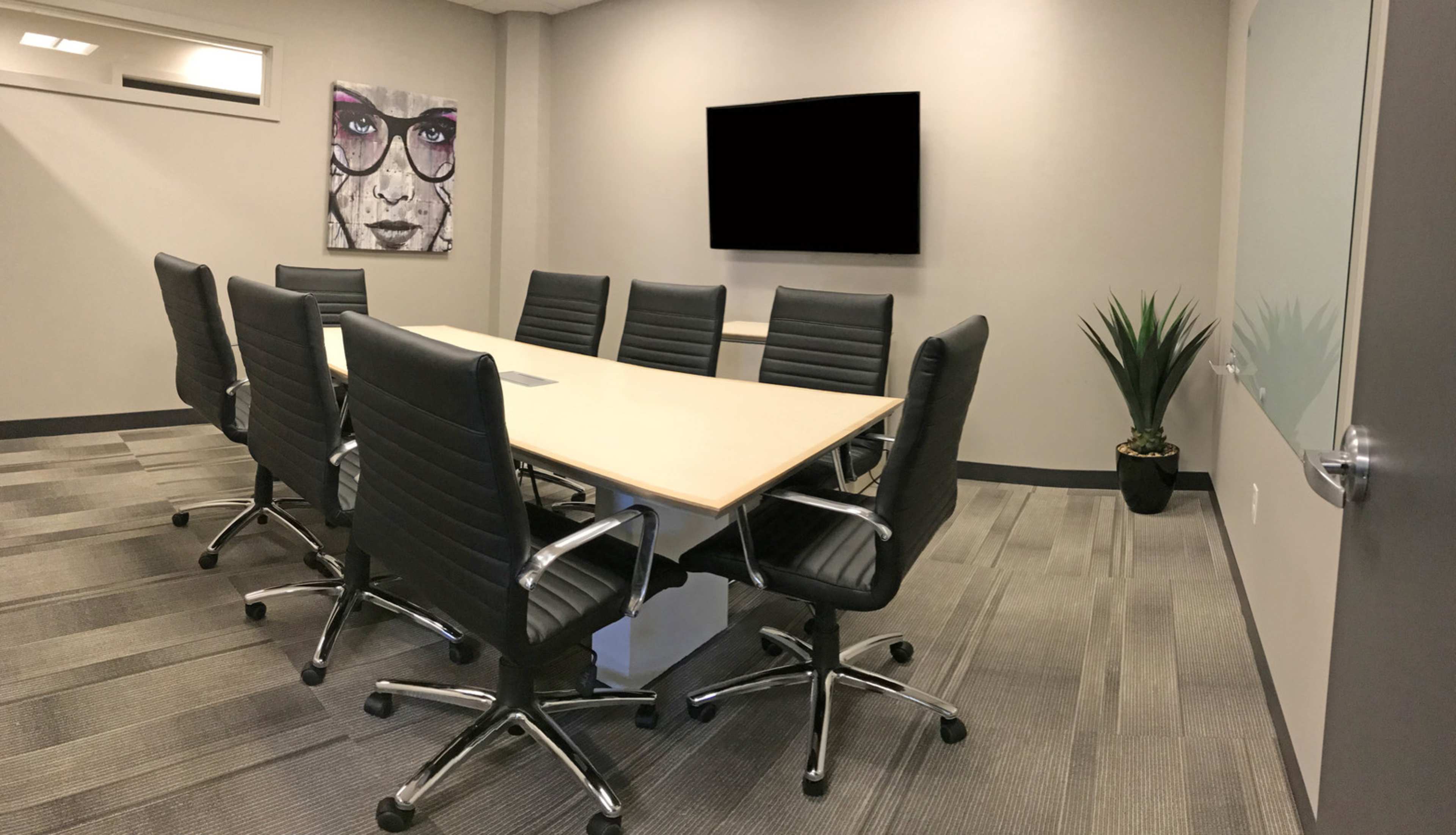 A modern conference room with a large table surrounded by black office chairs, a television on the wall, and a decorative plant in the corner.