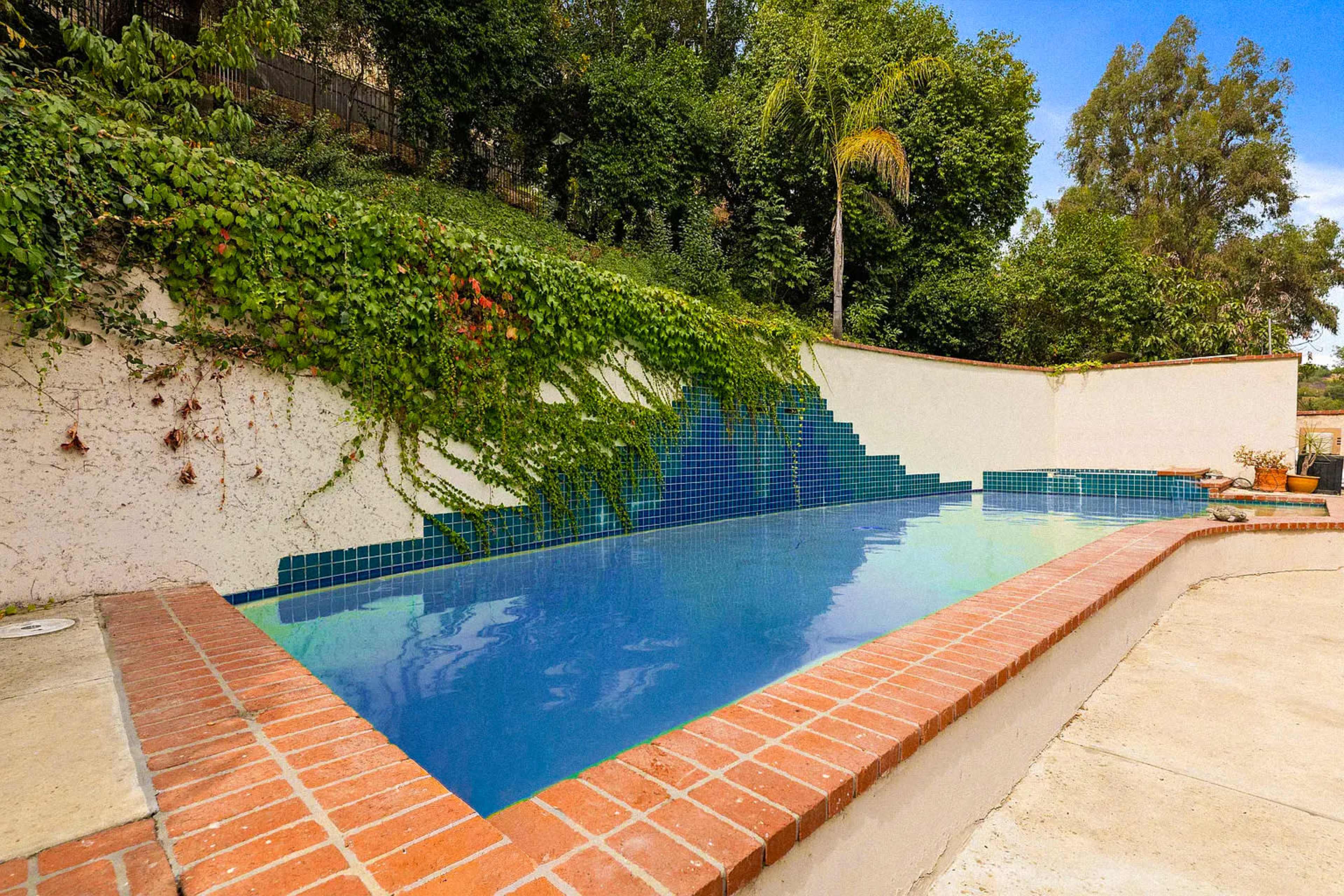 The image shows a swimming pool surrounded by brick coping, with a backdrop of lush greenery and a wall partially covered in ivy.