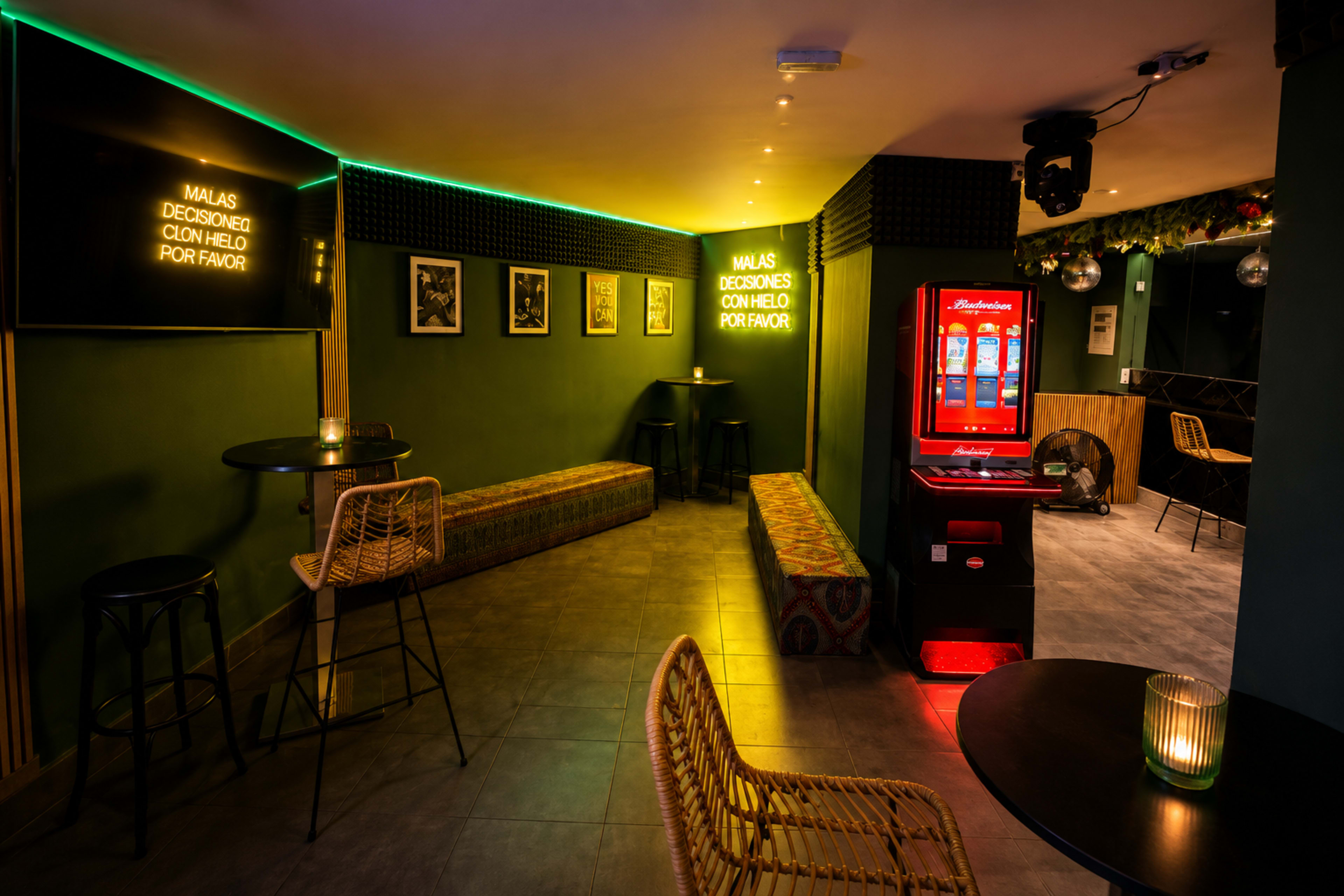 The image shows a dimly lit bar area with benches and tables, featuring neon lights, framed pictures, and a vending machine against a green wall.