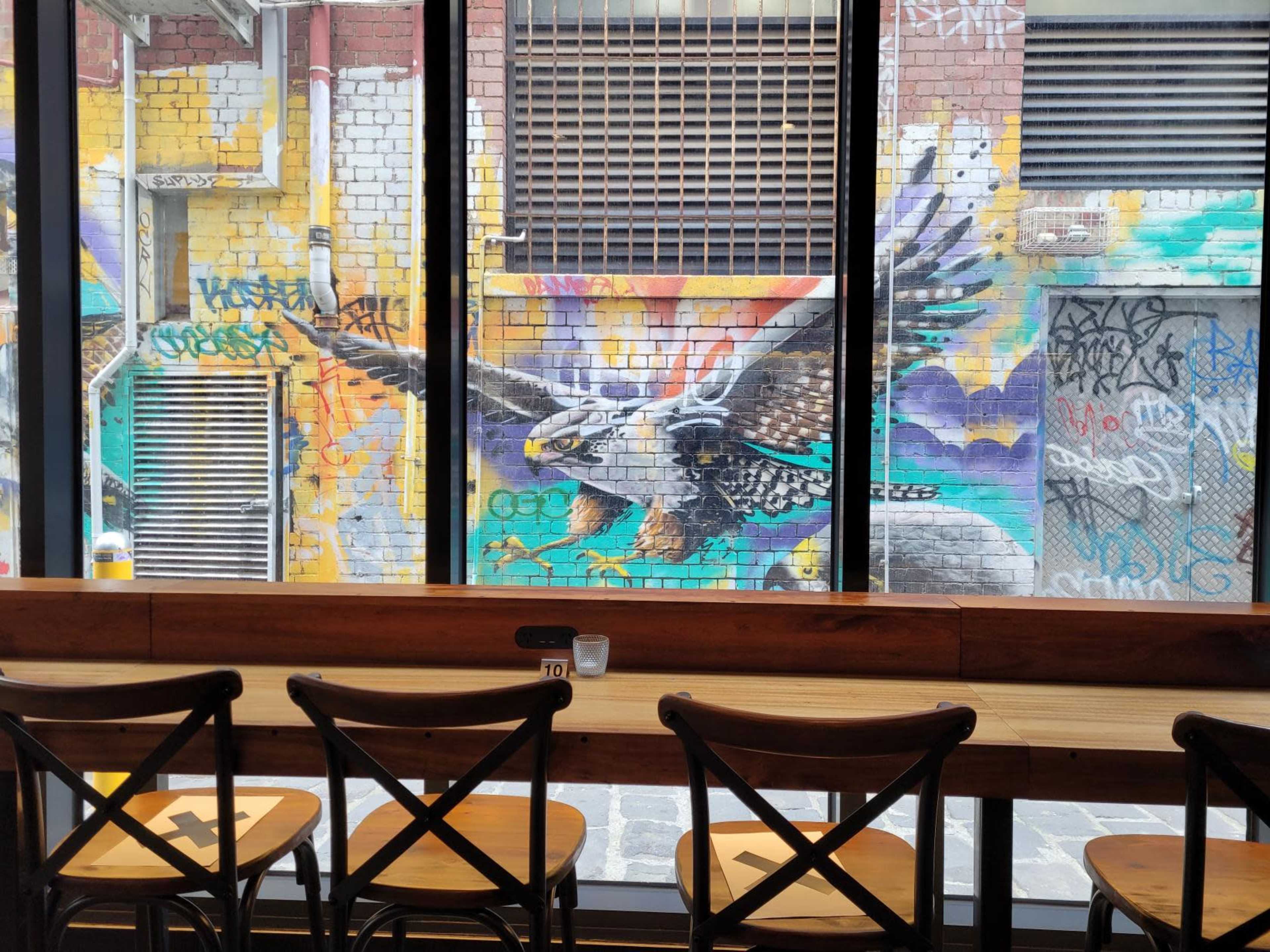 A mural of an eagle on a colorful brick wall, viewed through large windows from inside a café with wooden seating.