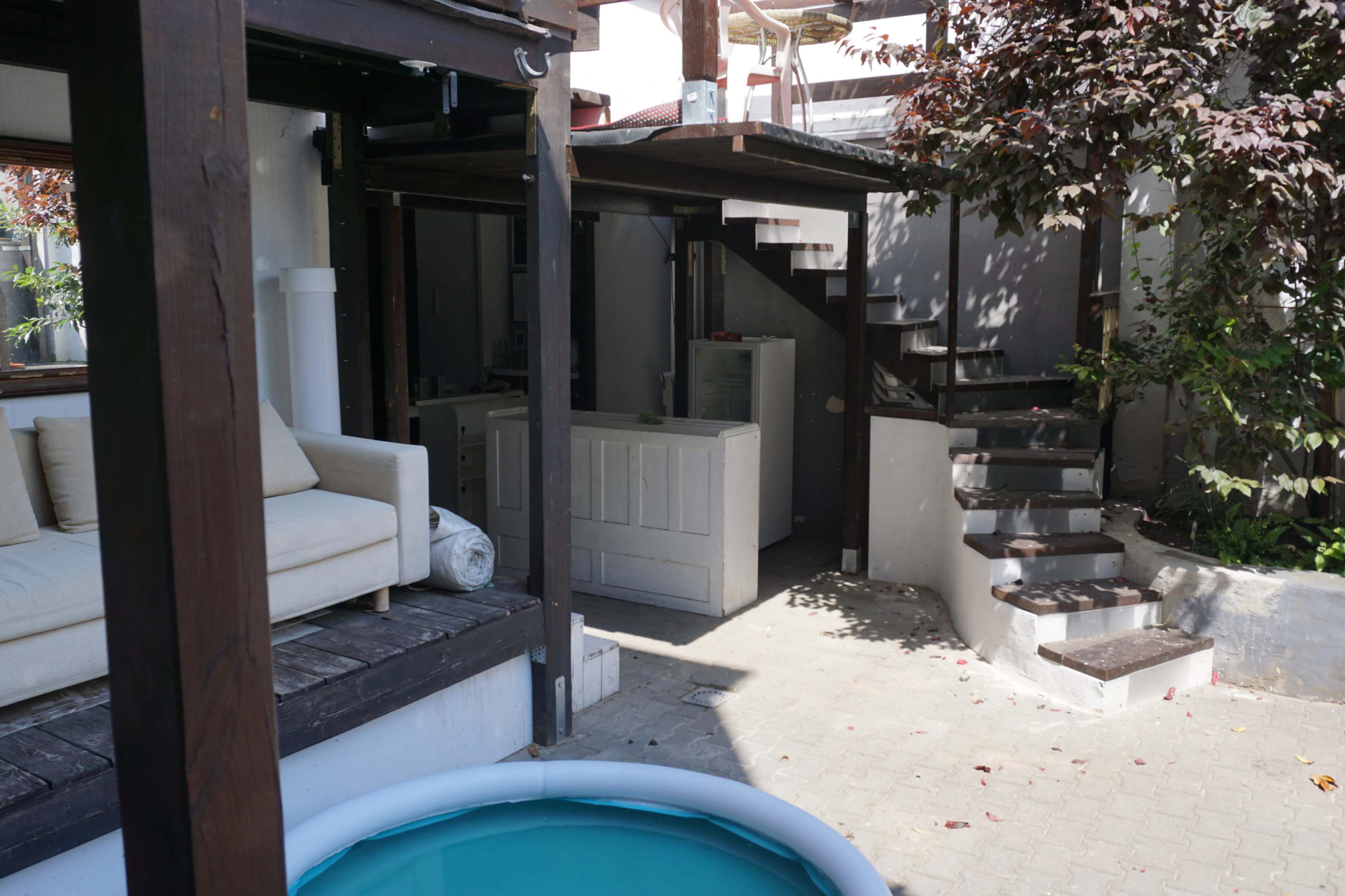The scene features a raised wooden deck with a sofa near a small pool, adjacent to a staircase leading to a higher level and a kitchen area in an outdoor setting.