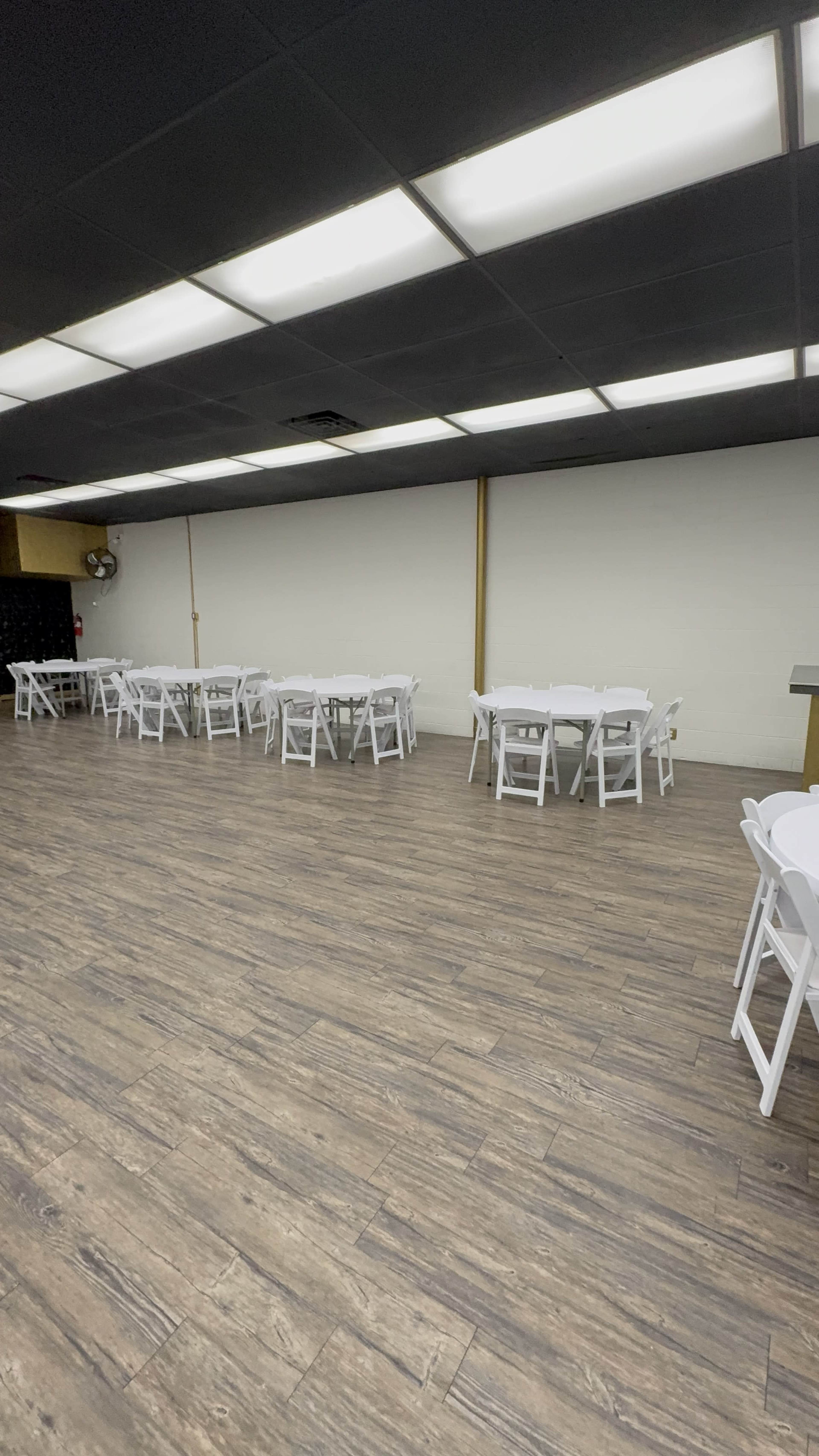 The image depicts a spacious room with several white tables and chairs arranged along the walls on a wooden floor.