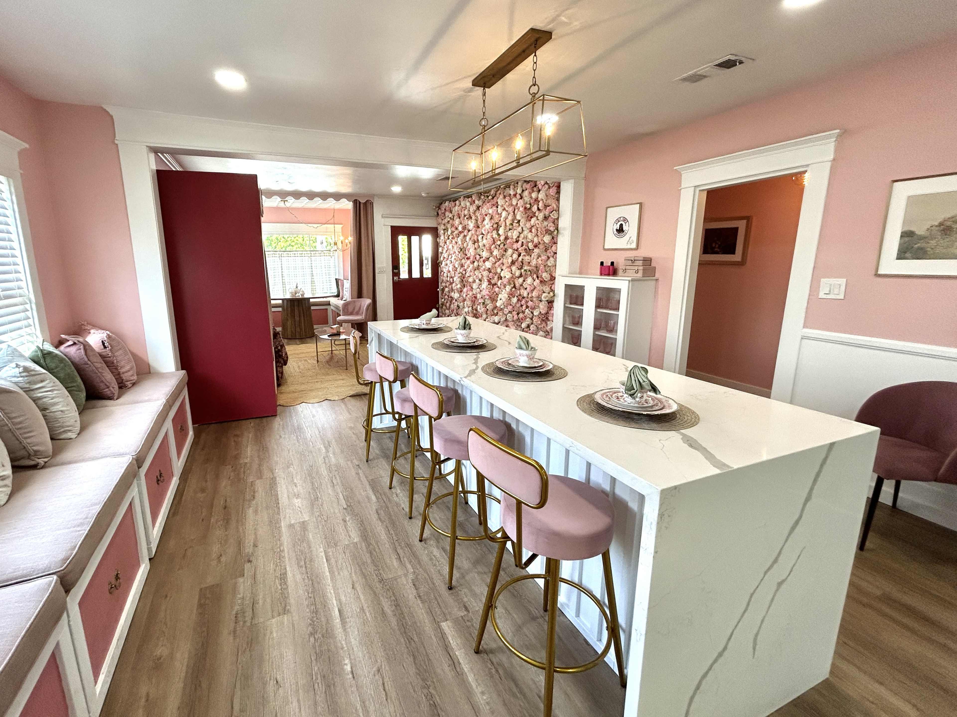 The image shows a bright dining area with a pink and white color scheme, featuring a long marble countertop with gold bar stools and floral wallpaper.