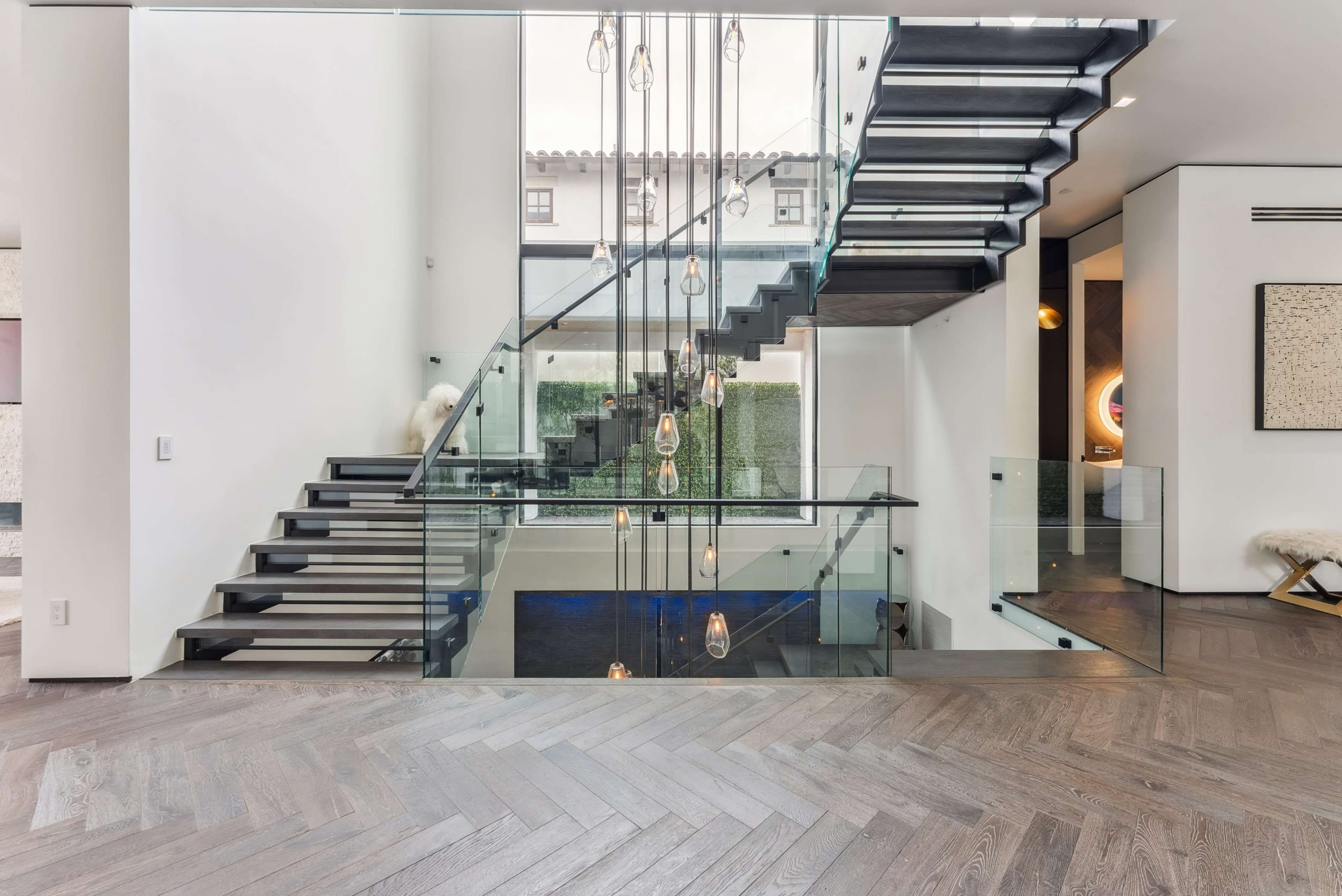 The image shows a modern interior with a glass staircase, wooden herringbone flooring, and pendant lighting.