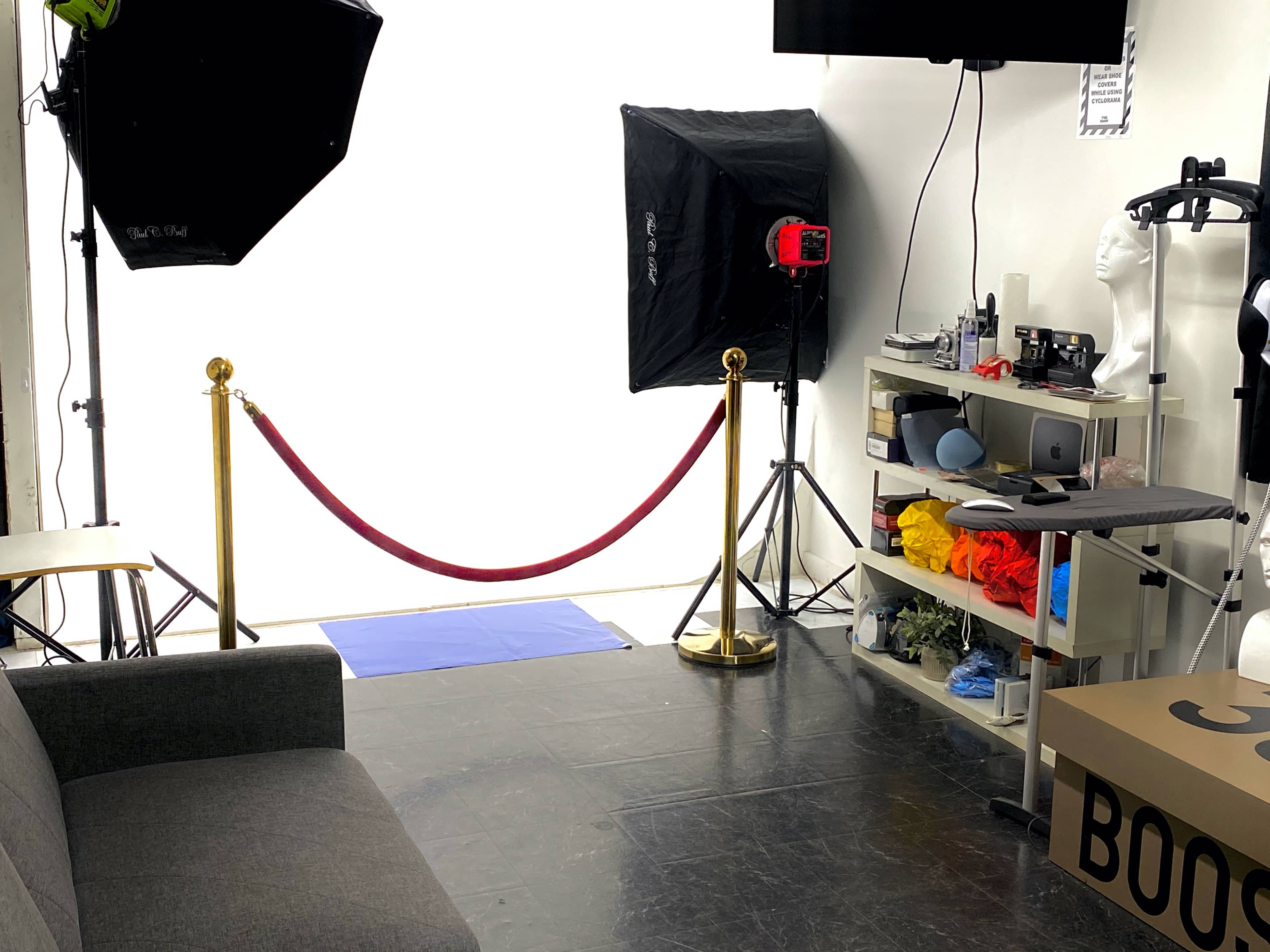 A studio setup with lighting equipment, a red velvet rope barrier, a gray couch, and various props on a shelf against a bright background.
