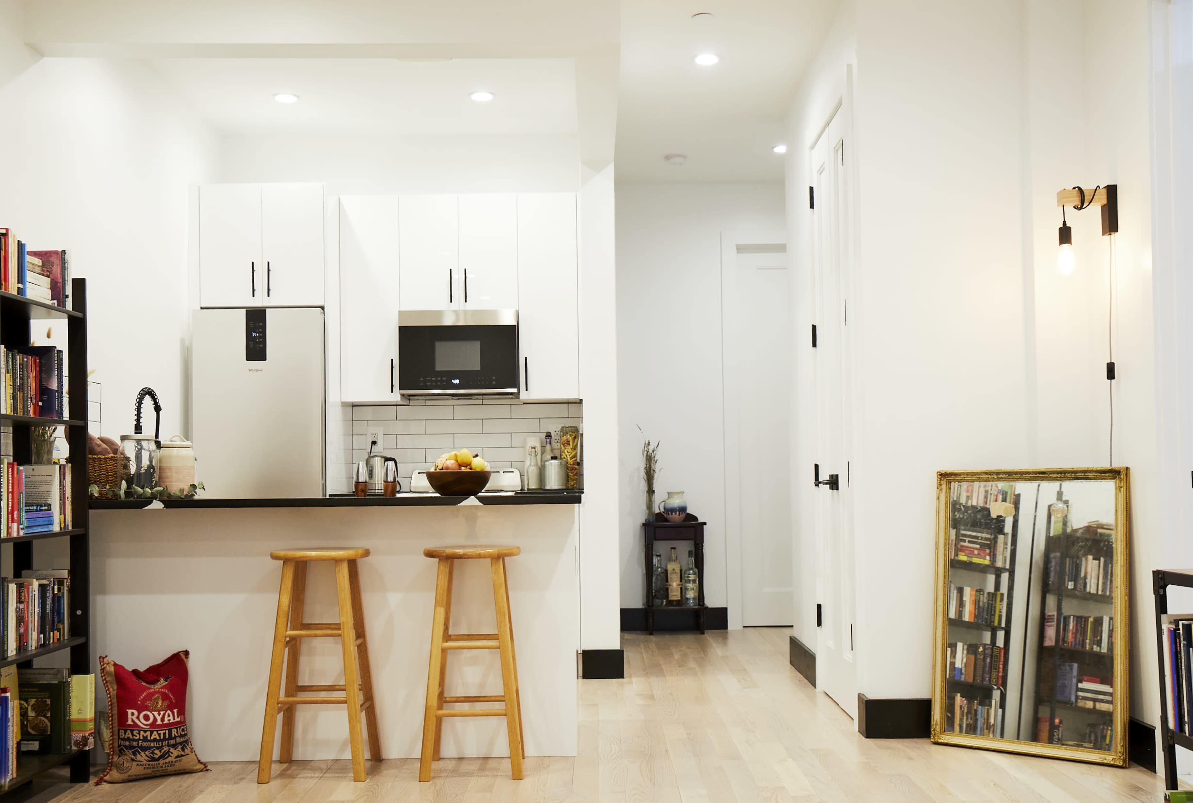 The image shows a modern kitchen and living space with a black countertop, two wooden stools, and a bookshelf filled with books, alongside a hallway leading to a door.