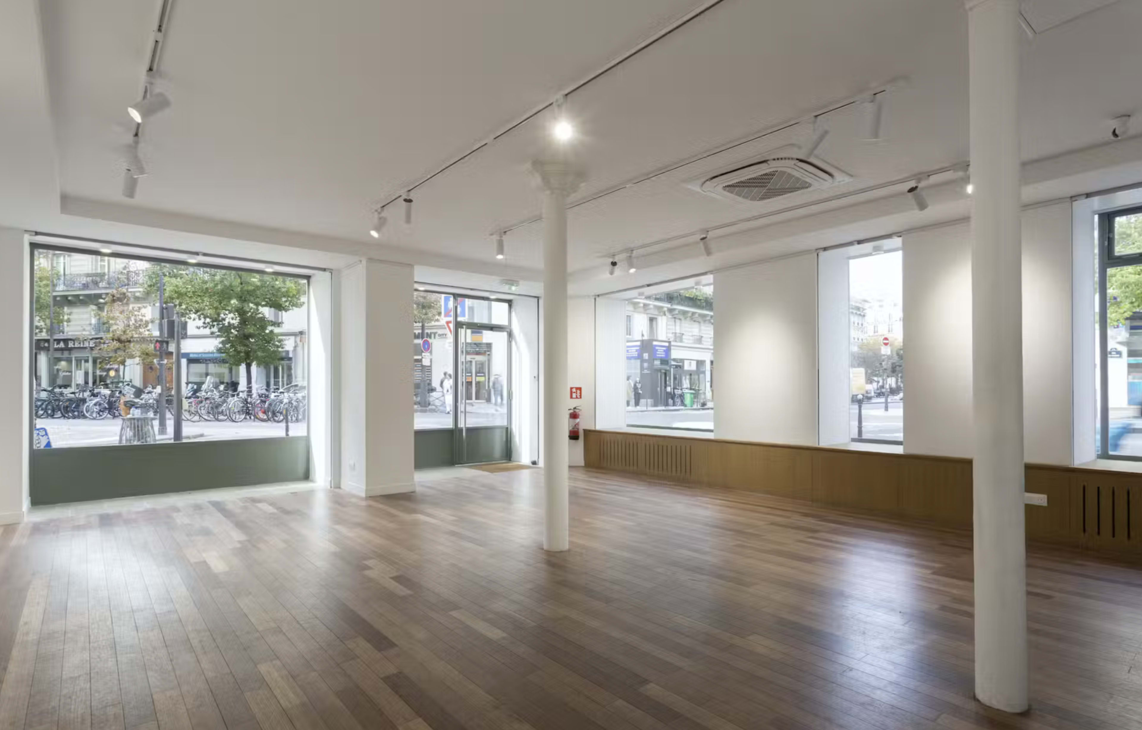 The image shows an empty gallery space with wooden flooring and large windows that overlook a city street.