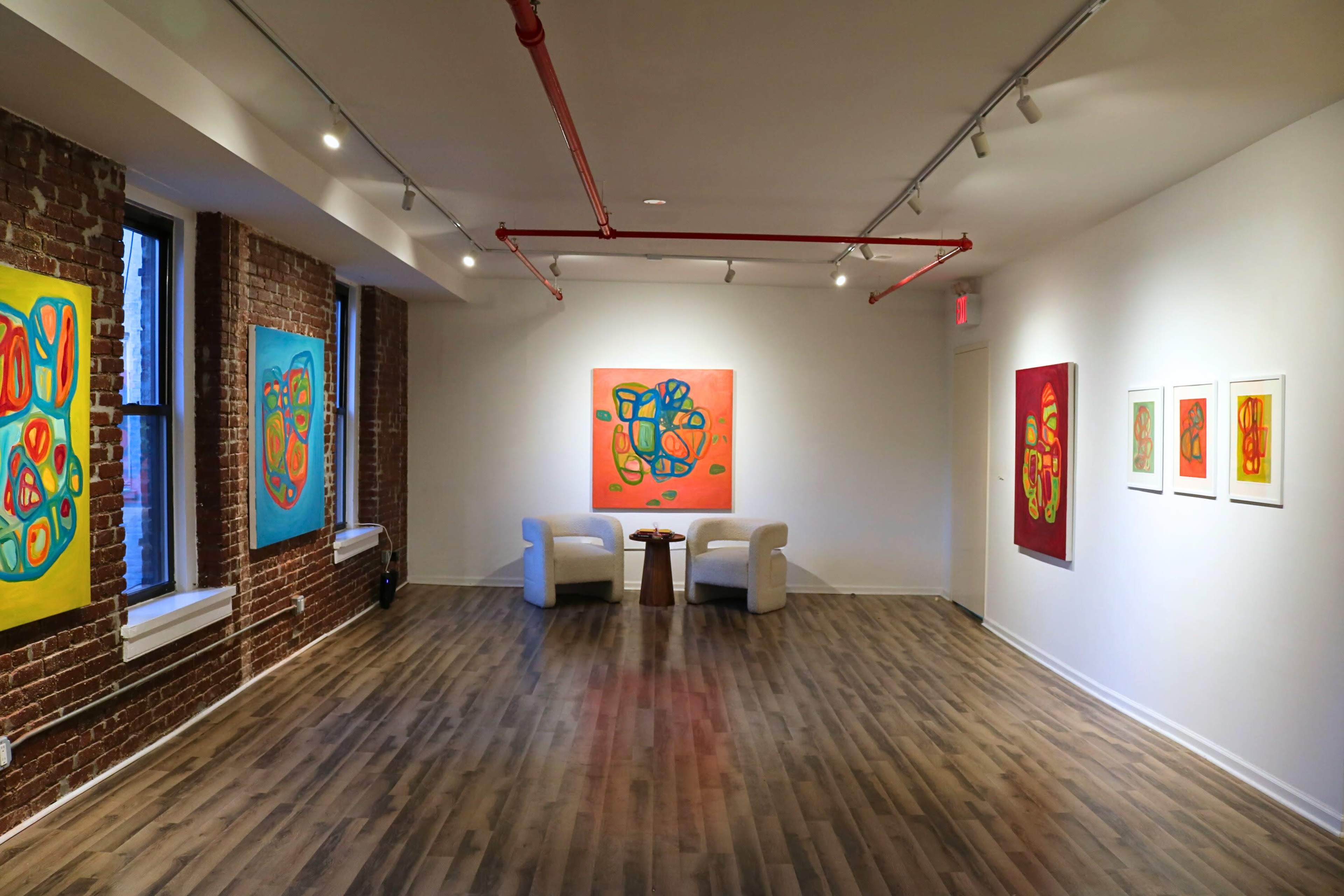 The image shows an art gallery with brick walls and modern paintings displayed on white walls, featuring two white chairs and a small table in the center.