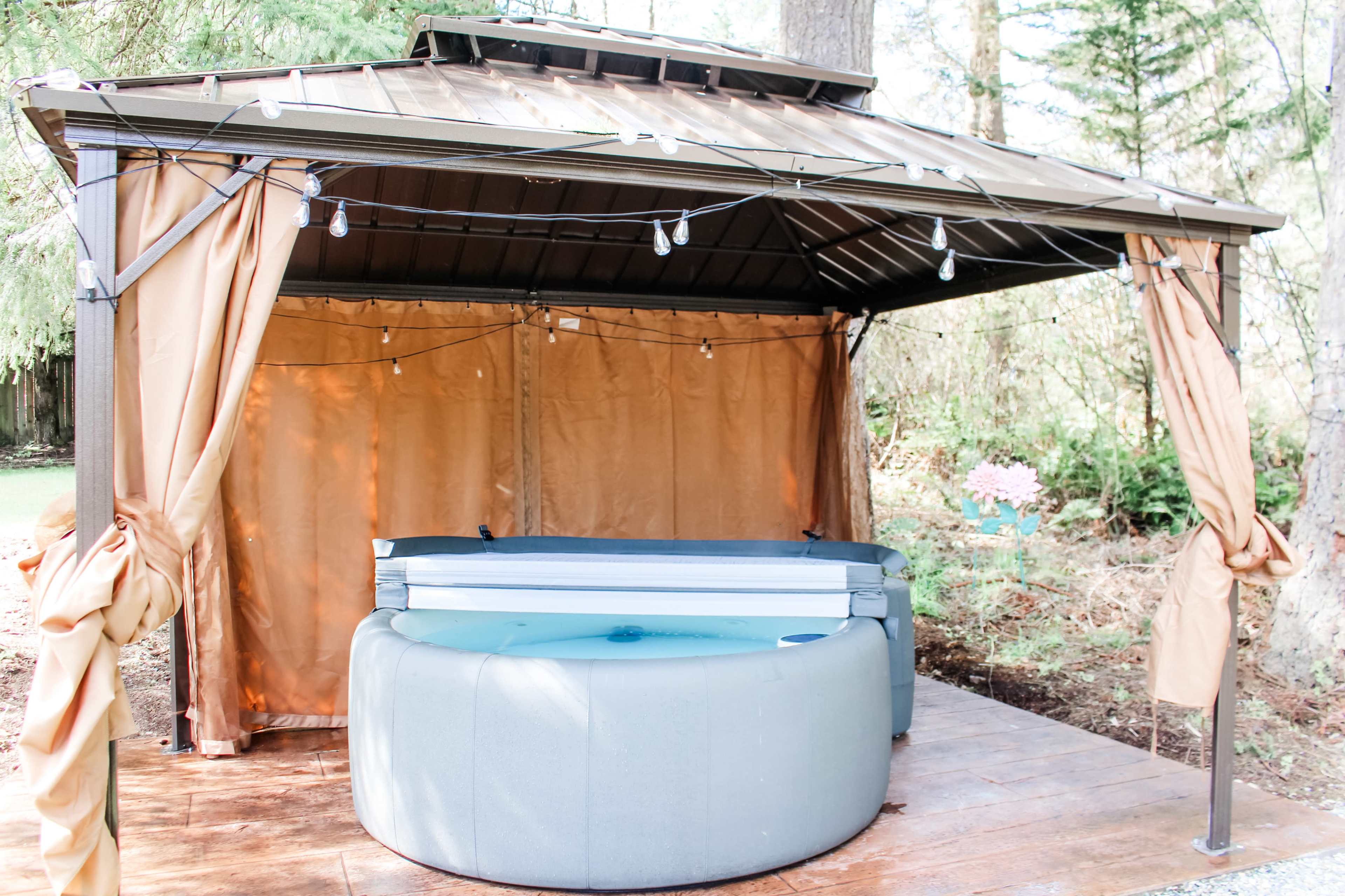 A shaded gazebo with curtain sides houses a hot tub under string lights in a wooded outdoor setting.