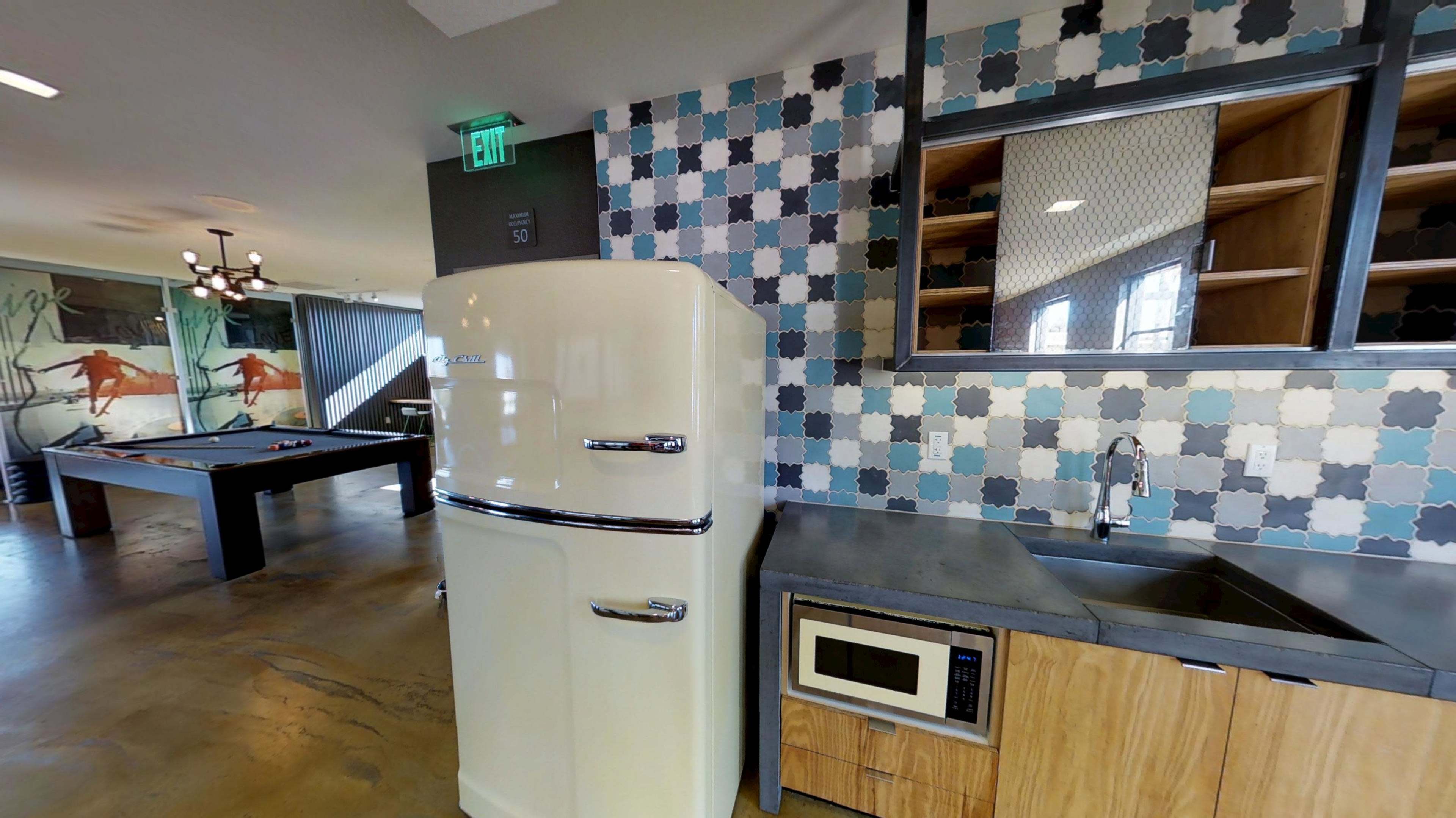 The image shows a vintage-style refrigerator next to a sink and microwave in a modern kitchen area with patterned tile walls and a pool table in the background.