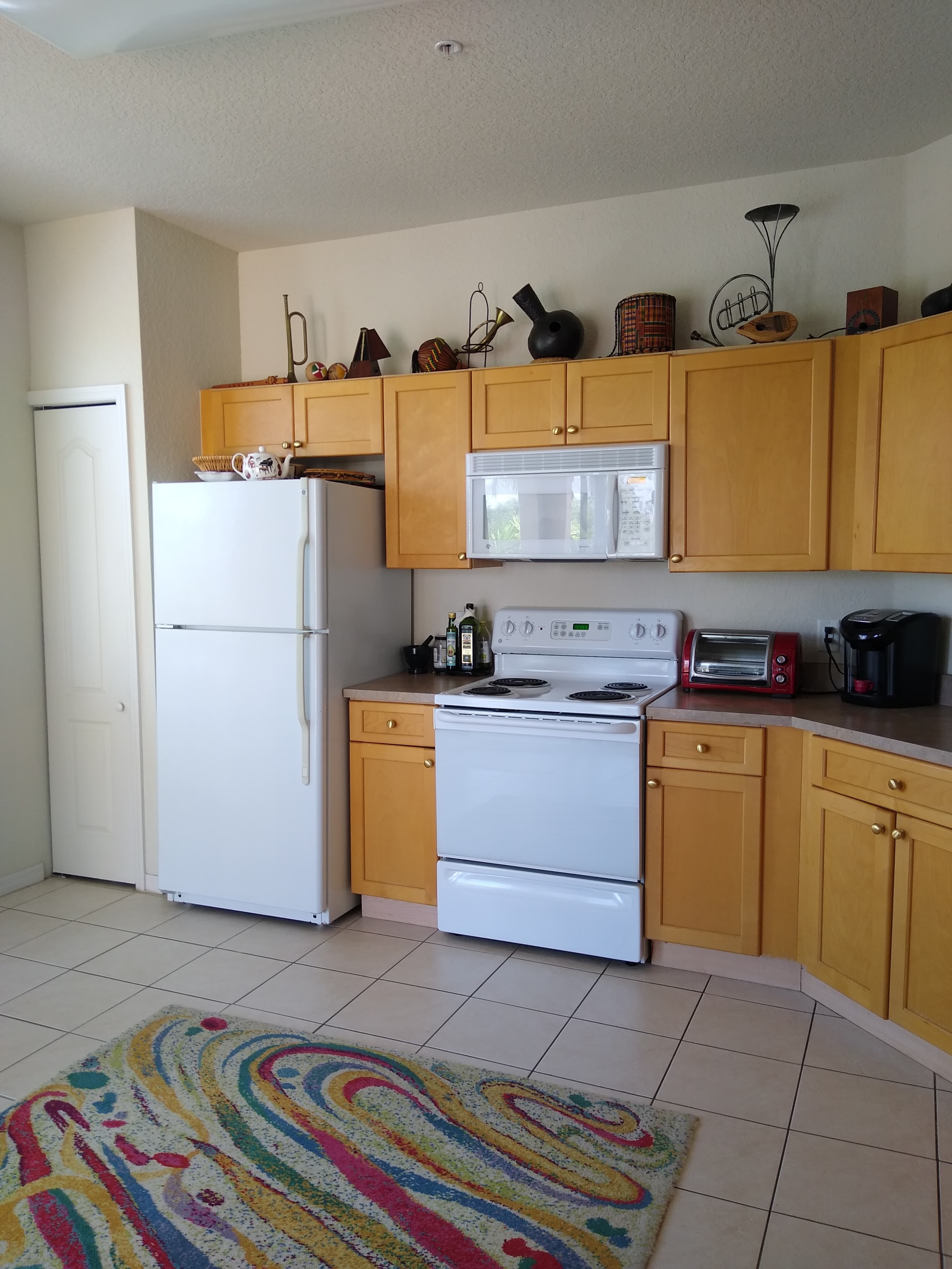 A kitchen features wooden cabinets, a white stove, a refrigerator, and various appliances with a colorful patterned rug on the tiled floor.