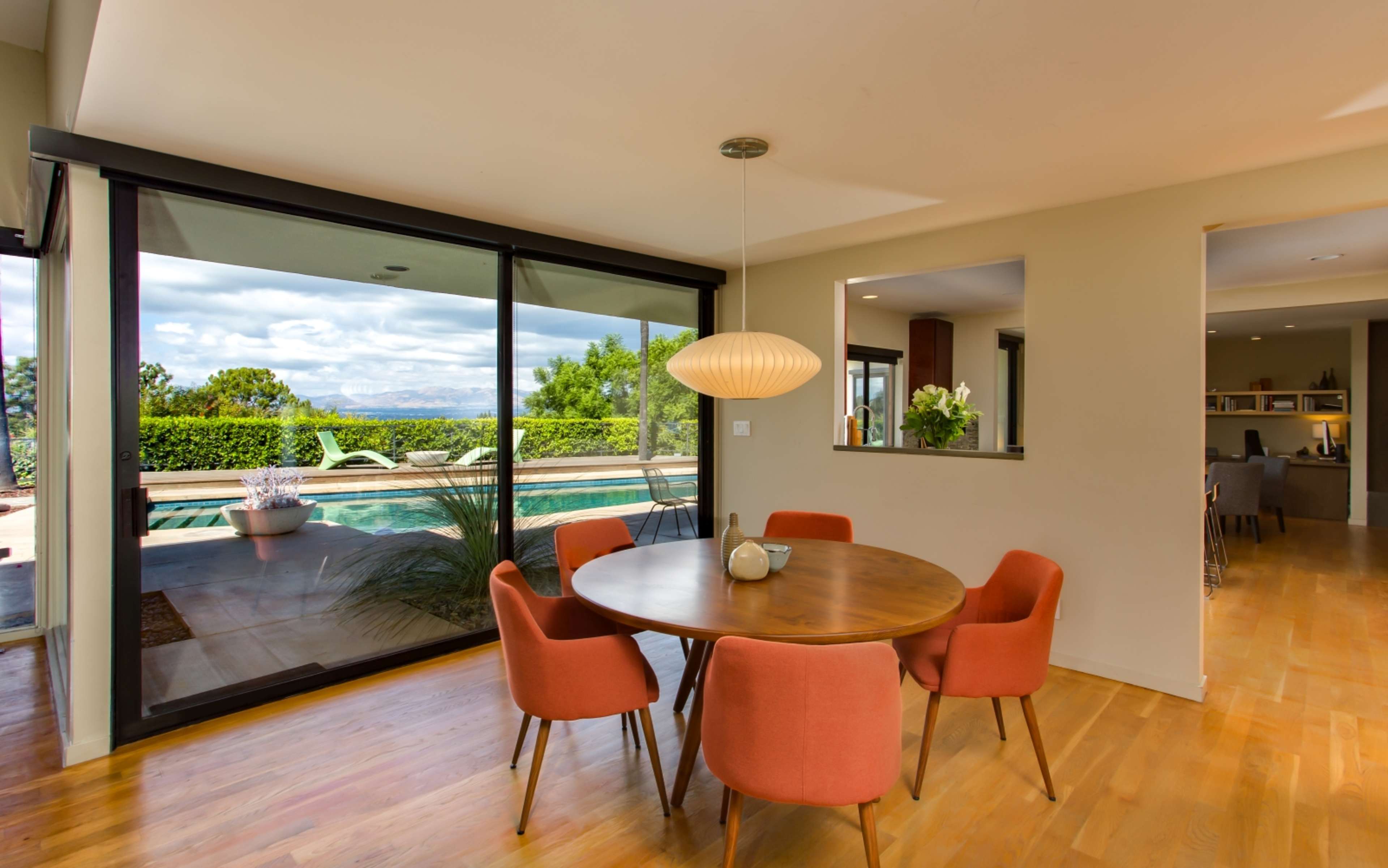 A dining area features a round wooden table surrounded by orange chairs, with large glass doors opening to a view of a pool and greenery outside.