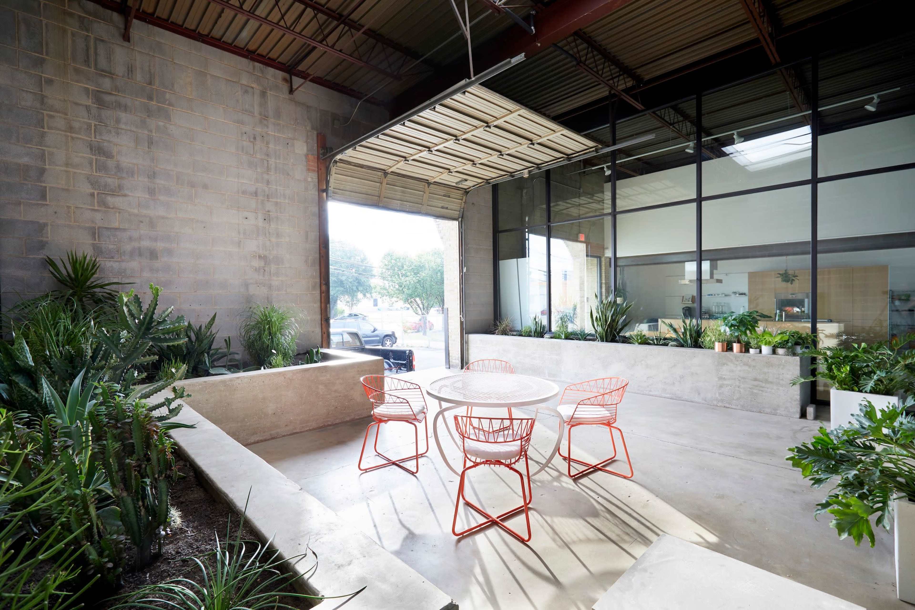 A modern outdoor seating area features a circular table surrounded by four red metal chairs, with concrete planters and lush greenery nearby.