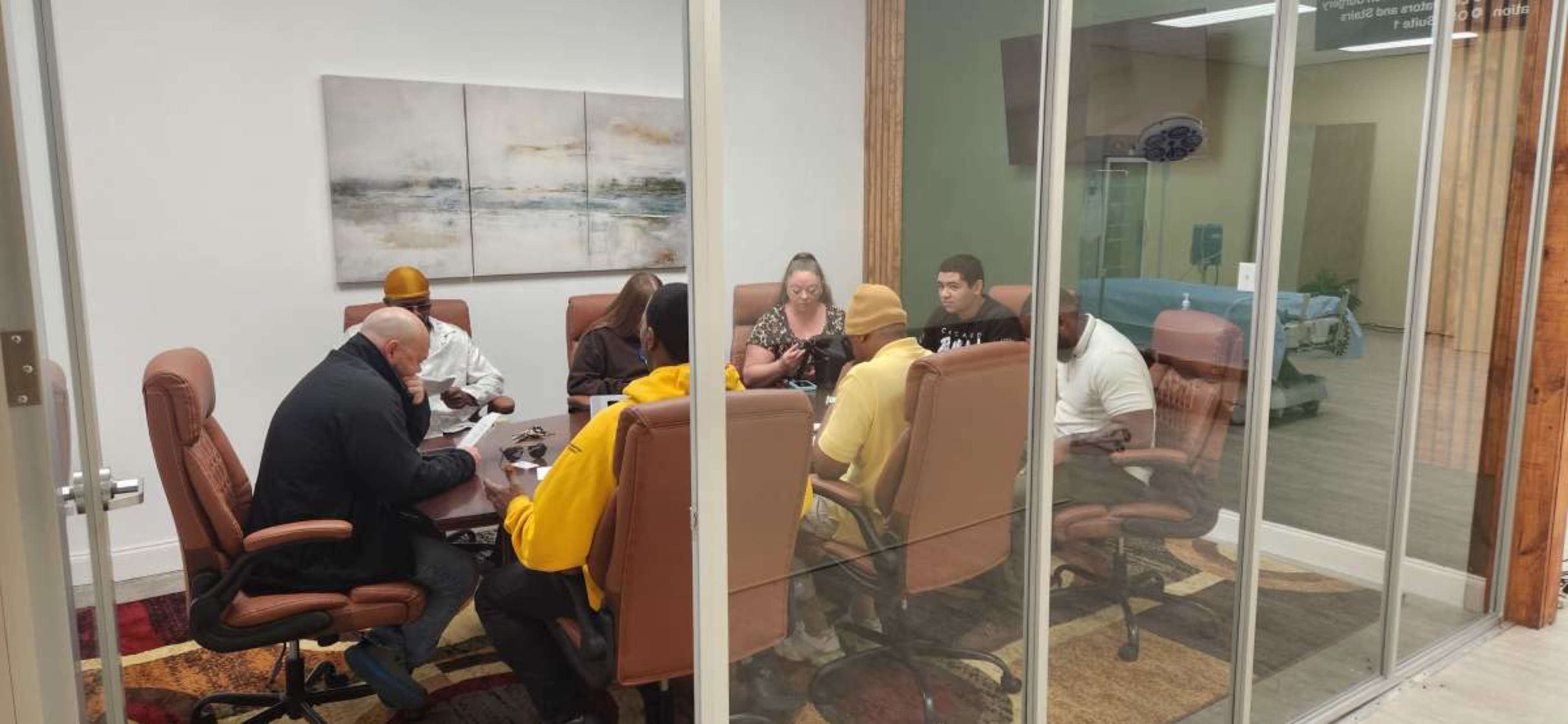 A group of eight people is seated around a conference table in a glass-walled meeting room.