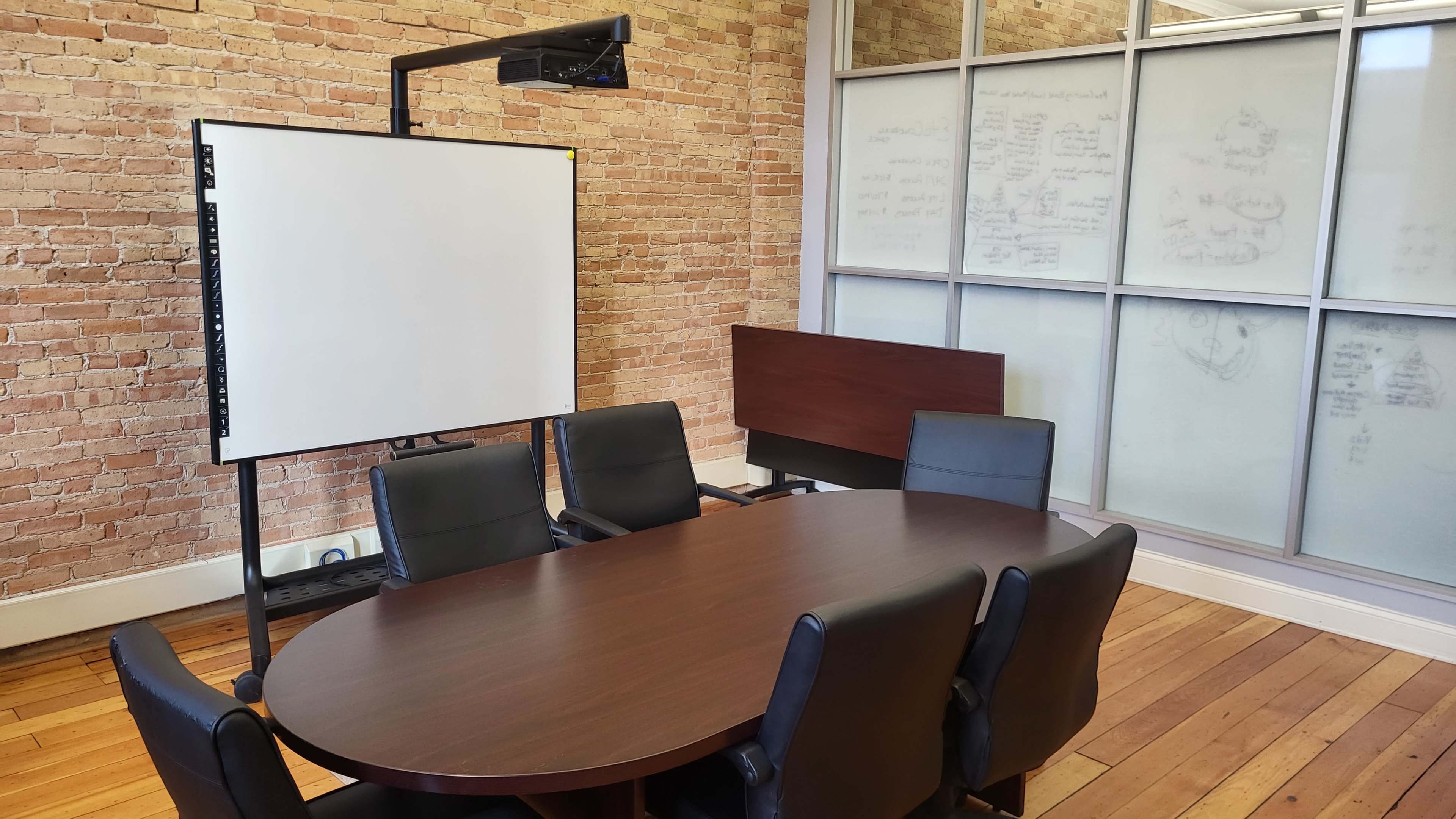 A conference room features a round wooden table surrounded by black leather chairs, with a whiteboard and a large glass wall displaying notes.