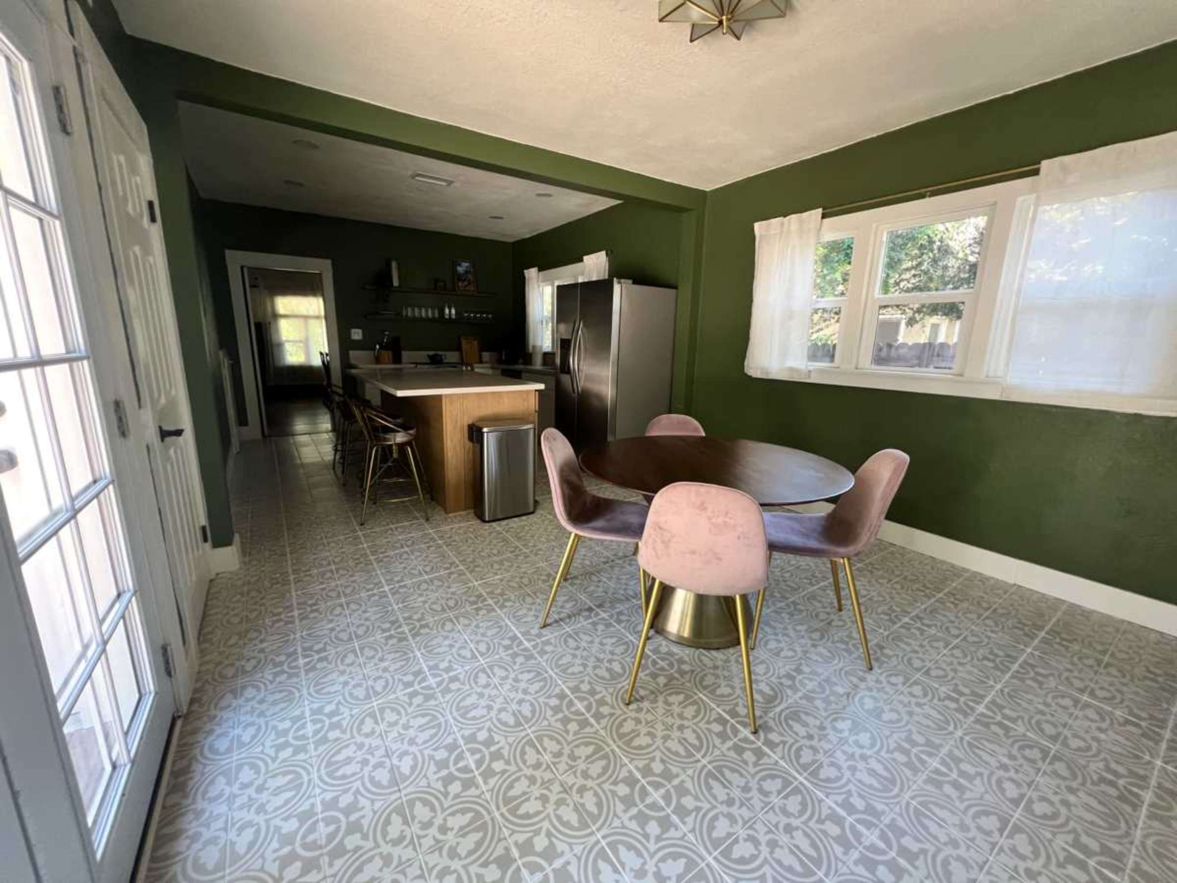 The image shows a modern kitchen with dark green walls, a round dining table with pink chairs, and stainless steel appliances.