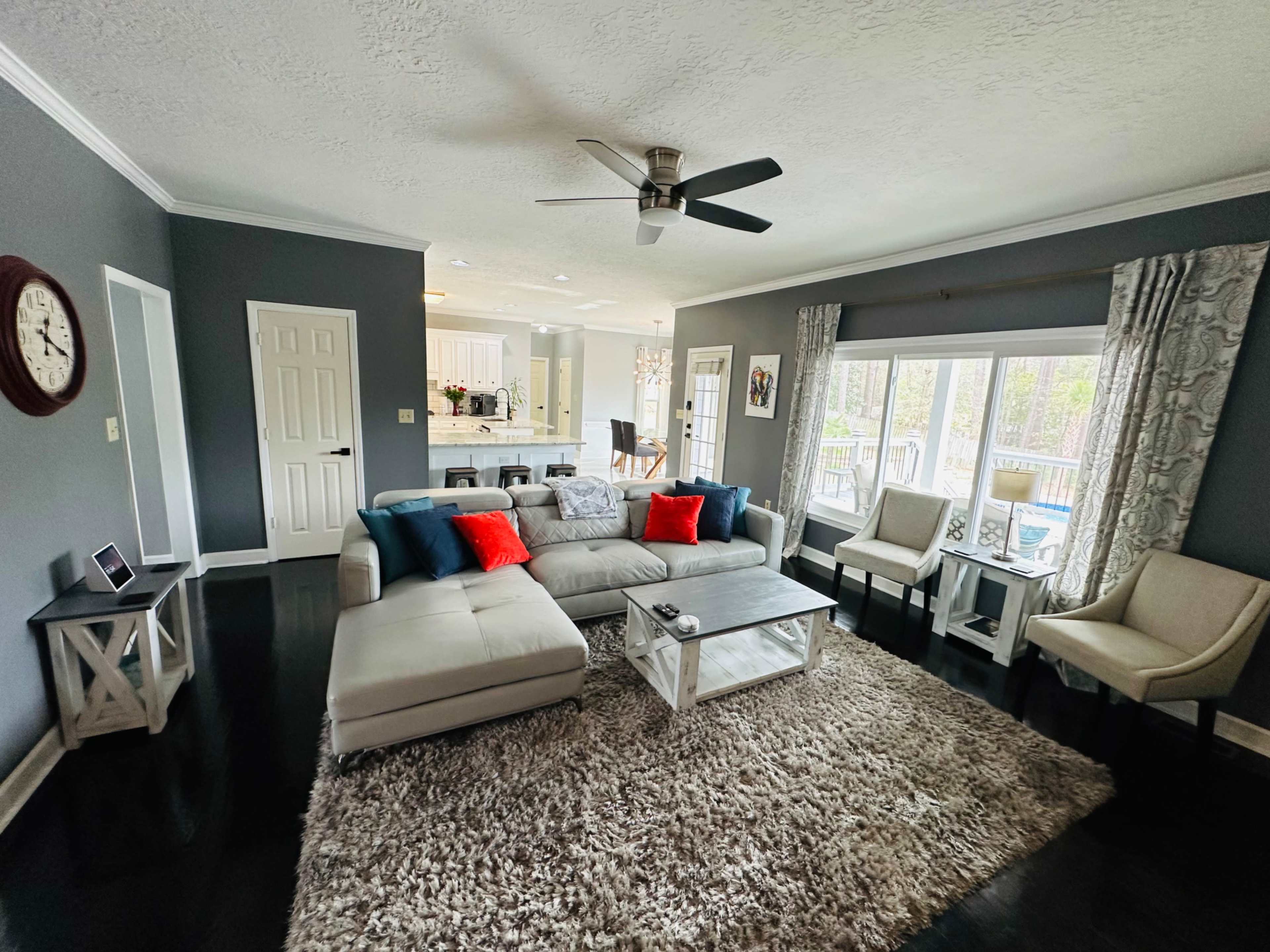 The living room features a gray color scheme with a sectional sofa, decorative pillows, a glass coffee table, and large windows allowing natural light to enter.