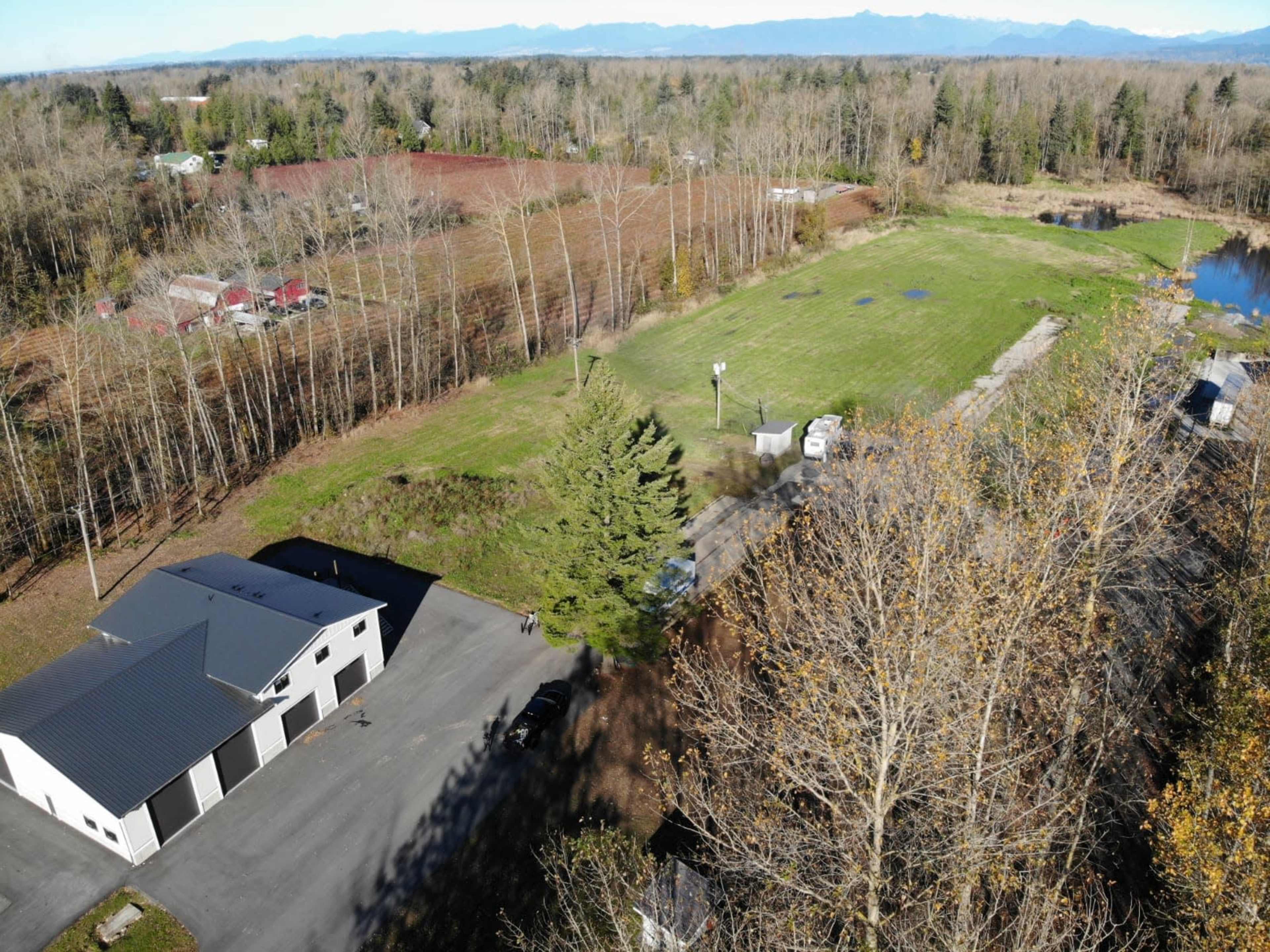 An aerial view of a rural landscape featuring a cleared field, scattered trees, a pond, and several buildings, with mountains in the background.