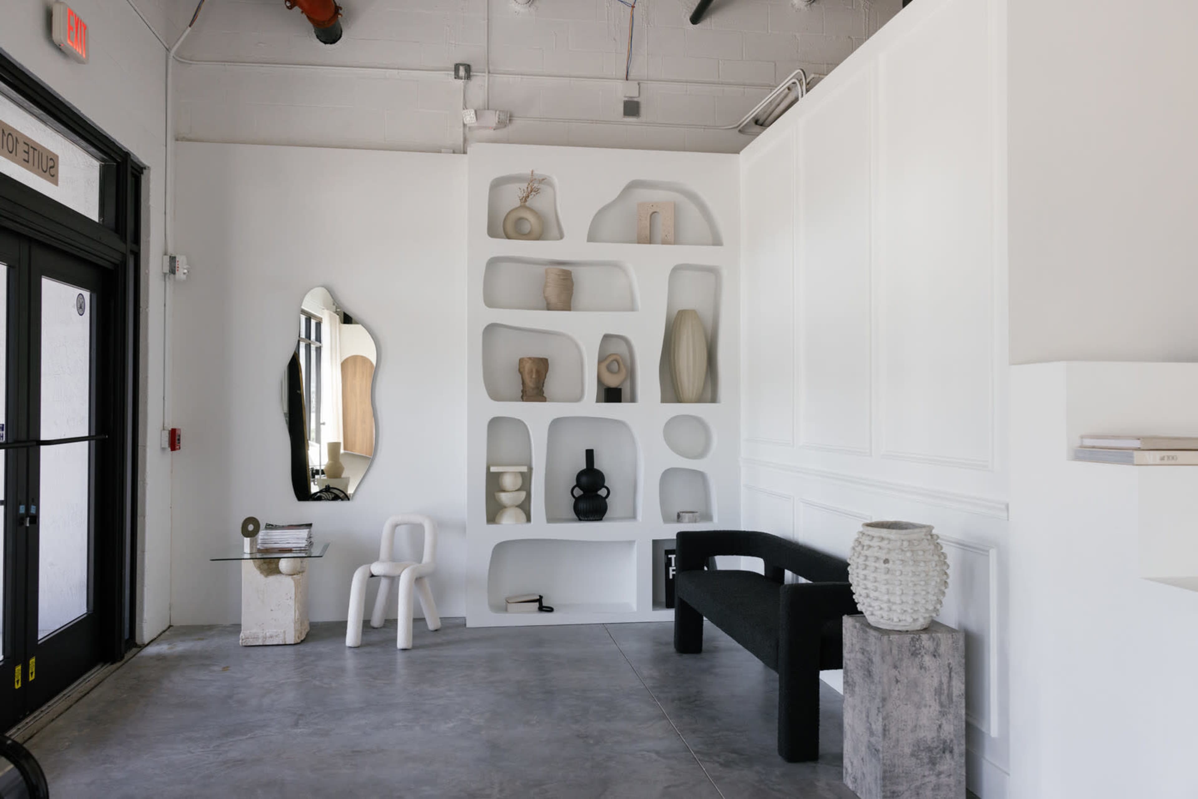 A minimalist interior featuring a sculptural wall display of various vases and objects, a black bench, a white chair, and a mirrored wall.