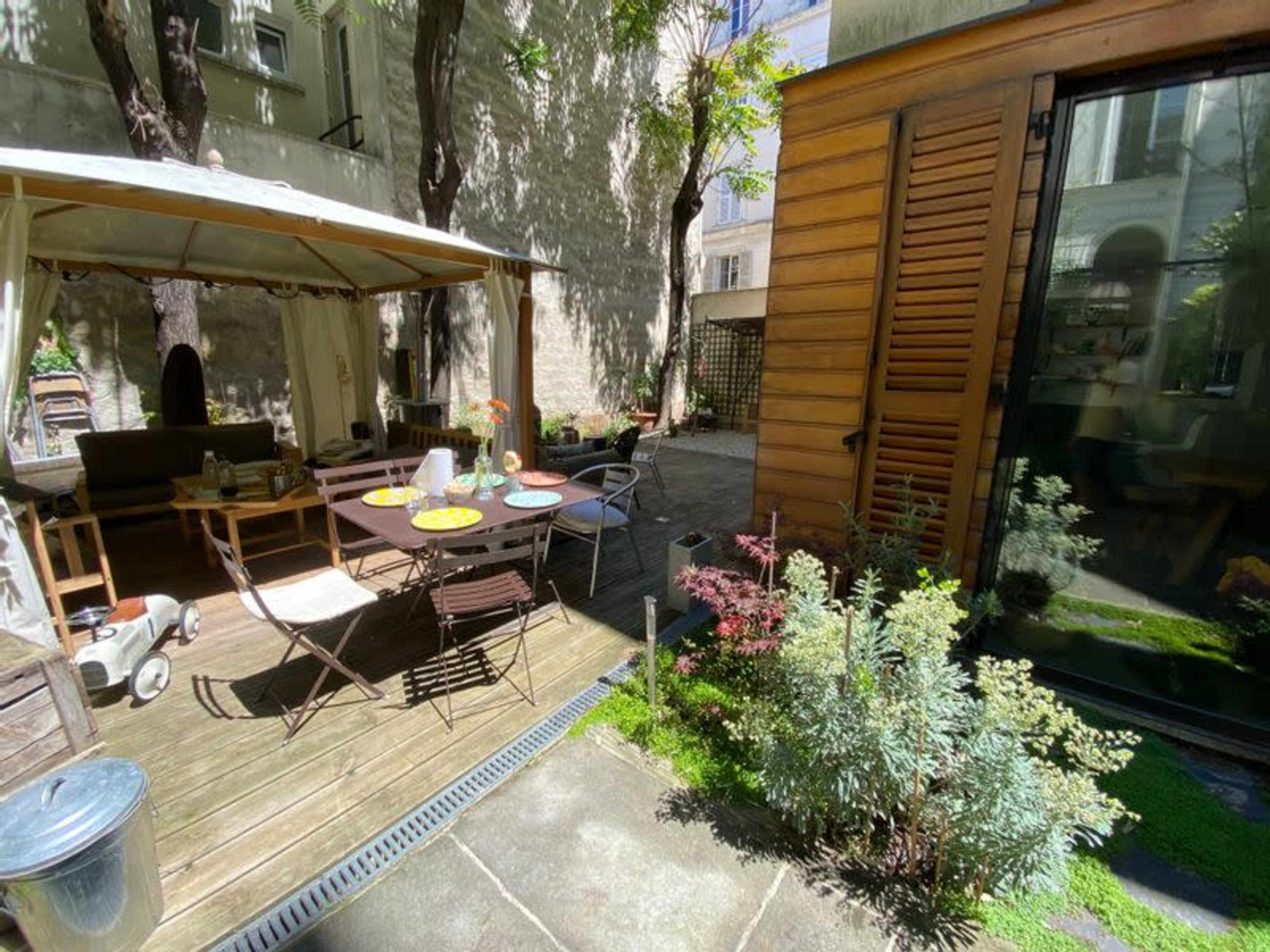 A small outdoor patio features a table set with dishes, surrounded by seating and greenery, adjacent to a wooden structure.