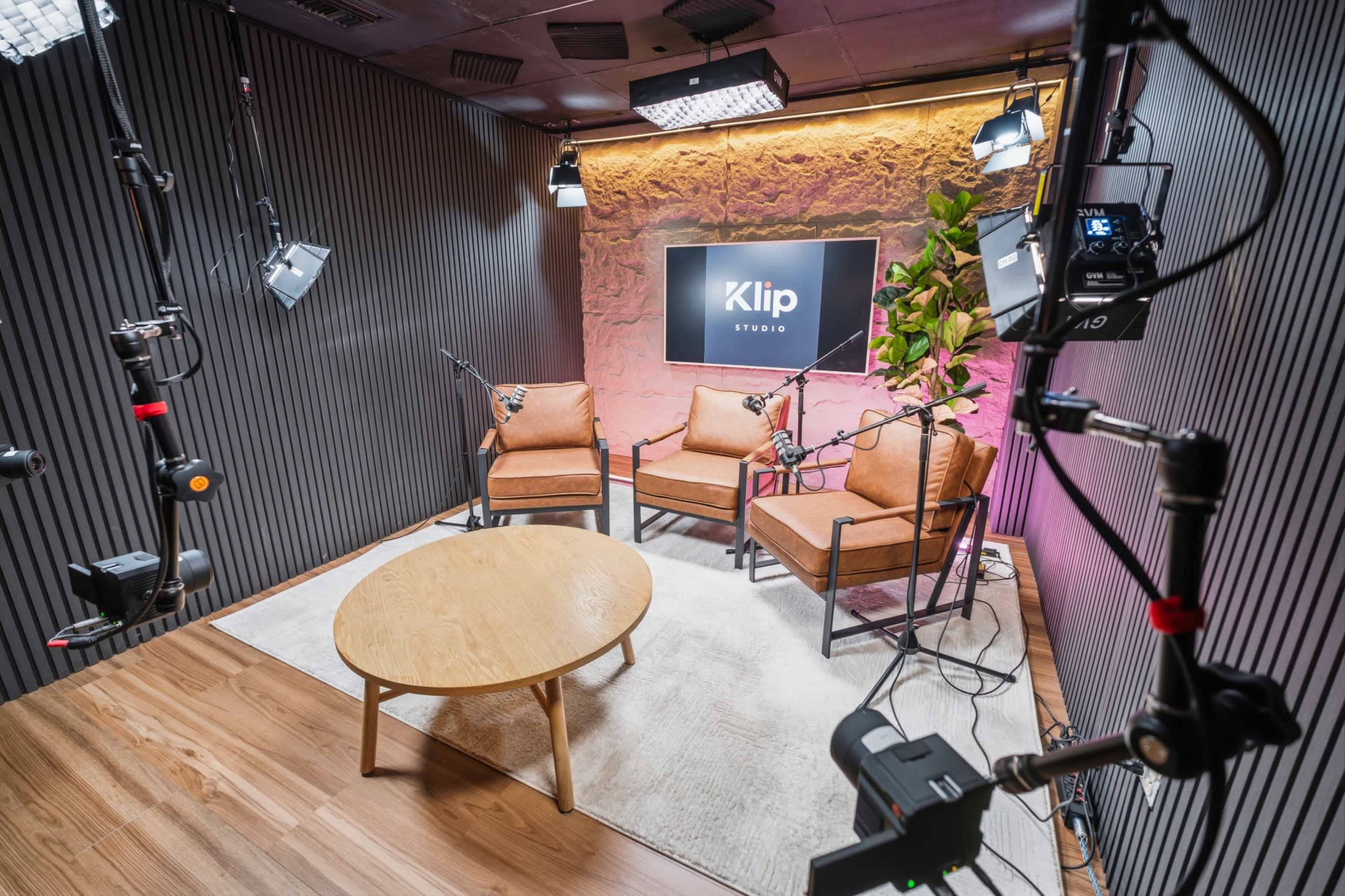 The image shows a modern studio setup with three chairs, a round wooden table, and various professional lighting equipment.
