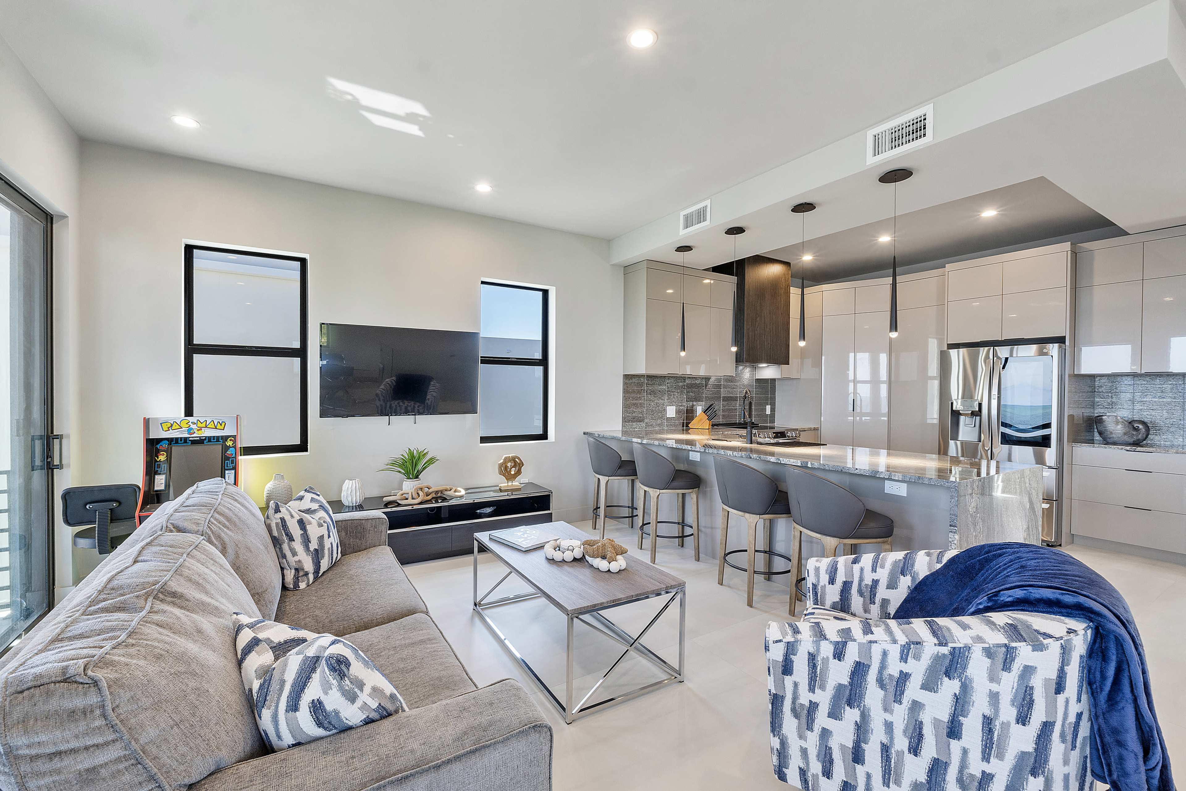 The image shows a modern living room with a gray sofa, a television mounted on the wall, and a kitchen area featuring sleek cabinets and bar stools.