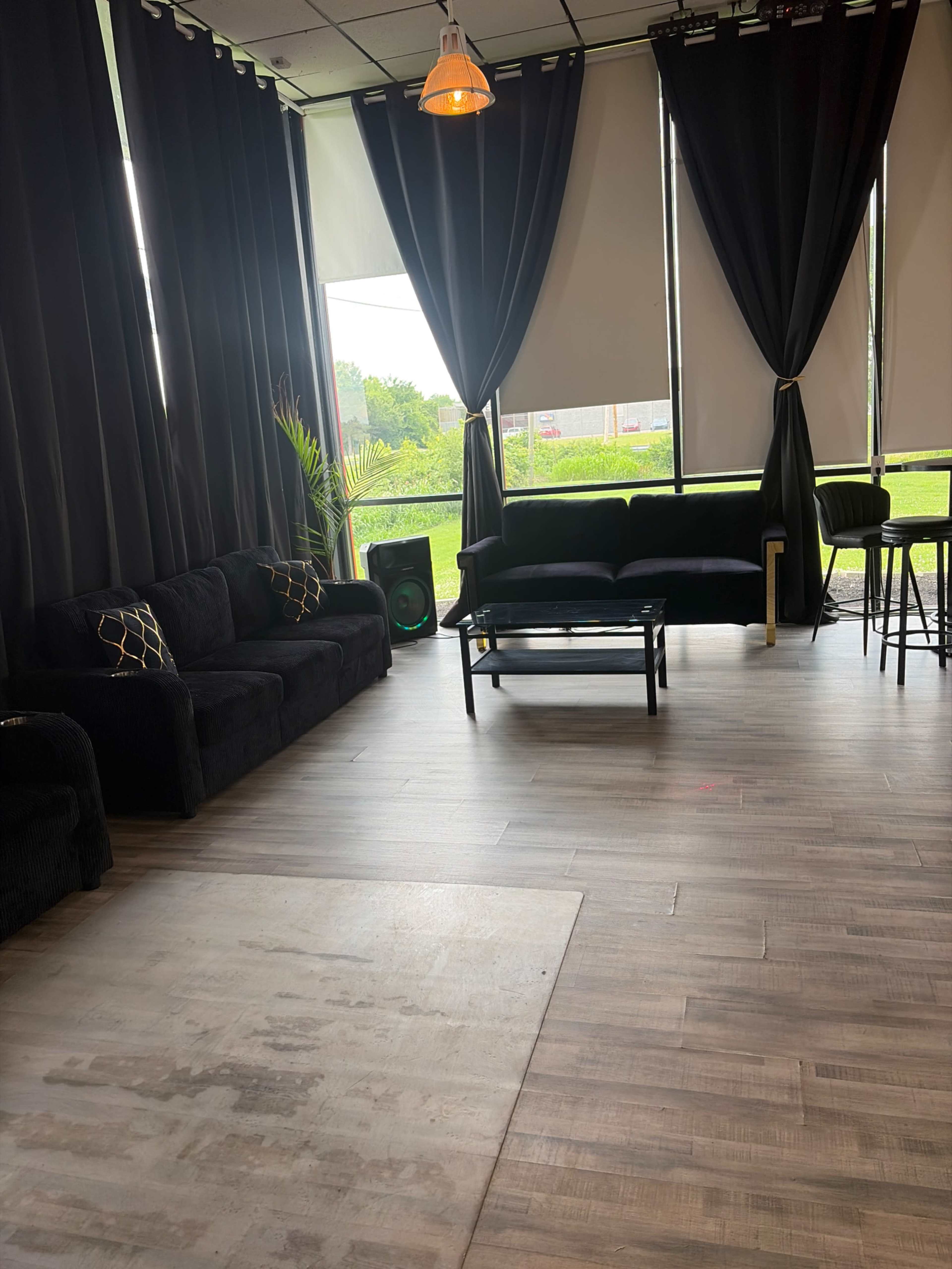 The image shows a modern interior space with dark curtains, two black couches, a coffee table, and a view of greenery through large windows.
