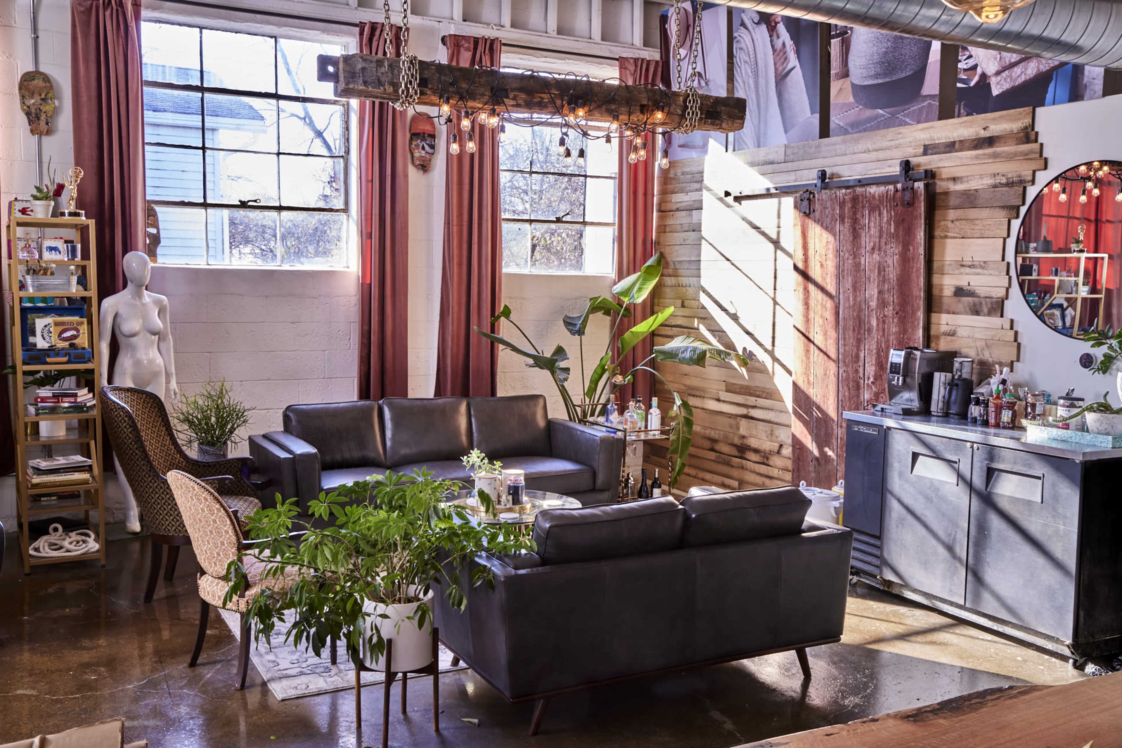 A modern living space with leather sofas, large windows, a wooden beam light fixture, potted plants, and a coffee station against a rustic wall.