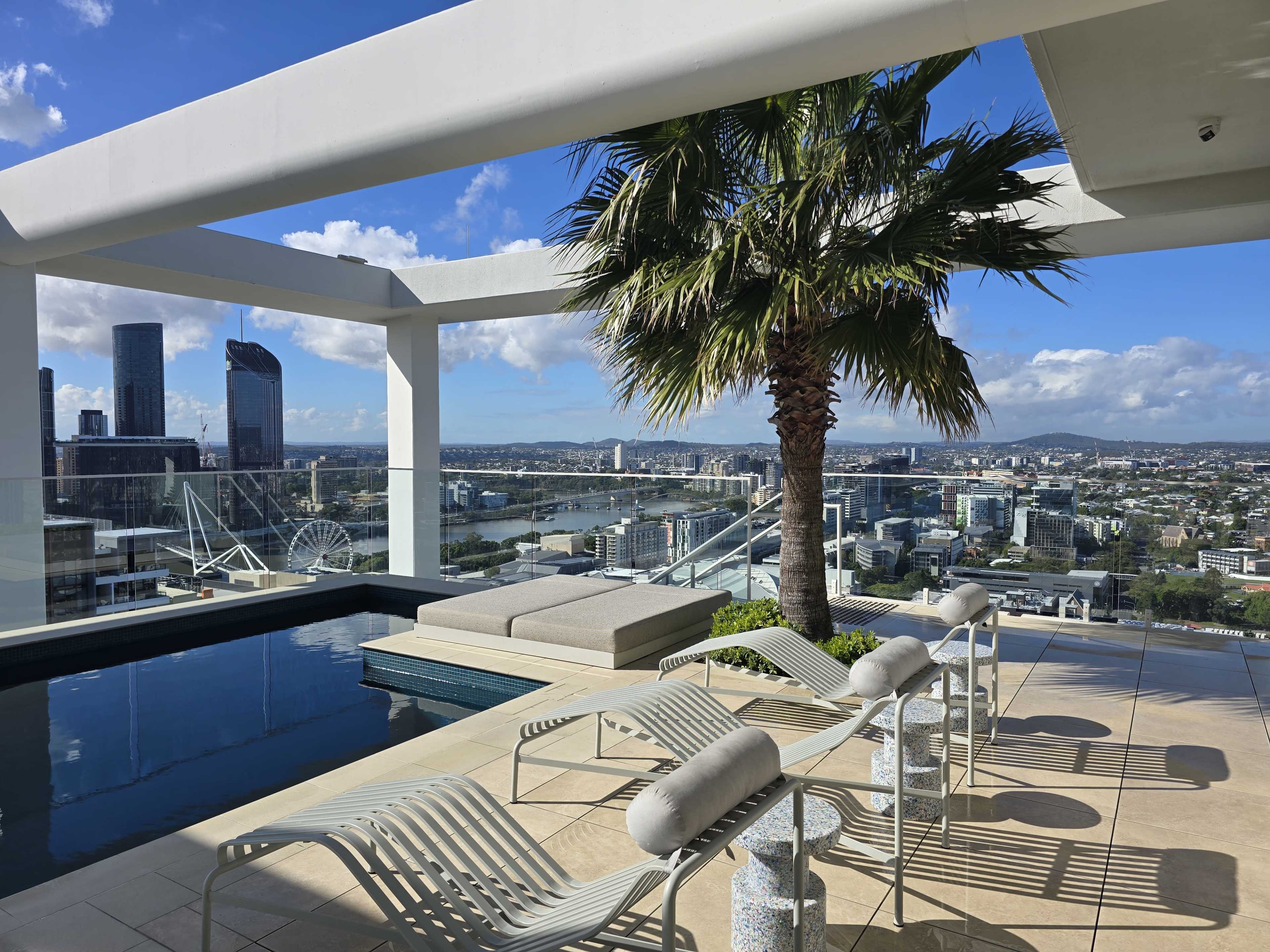 A rooftop pool area features lounge chairs and a palm tree, with a city skyline and river visible in the background.